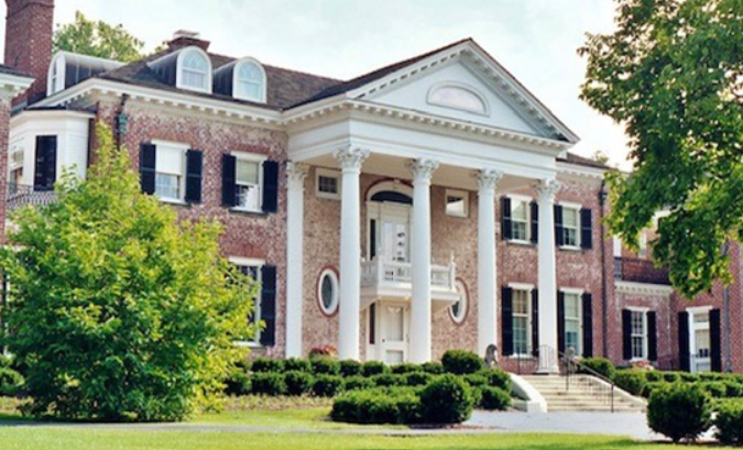 A two-story brick mansion with white columns, a large triangular pediment, and black shutters, set among trees and shrubs.