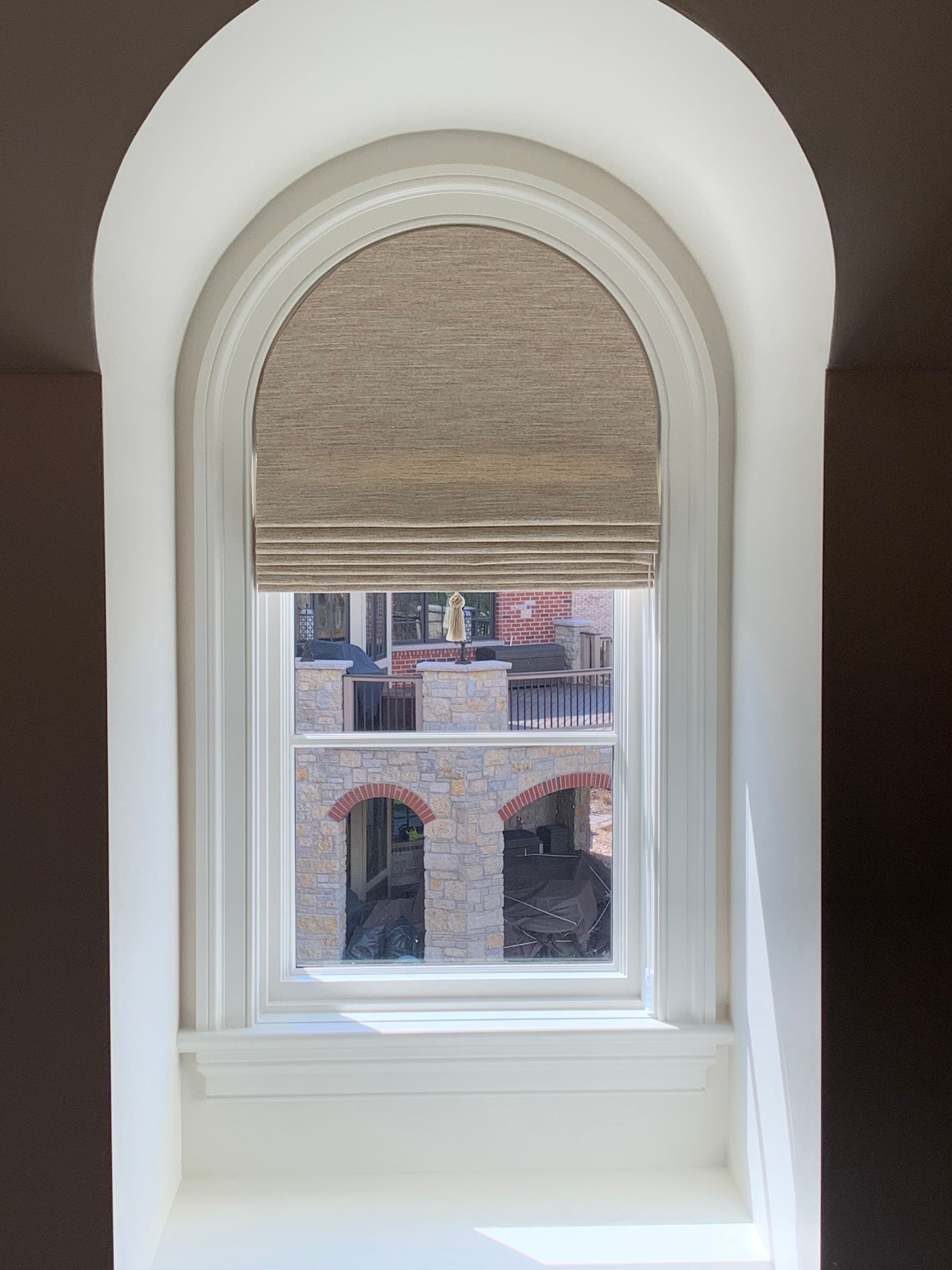 Arched window with beige Roman shade; view of brick building exterior.
