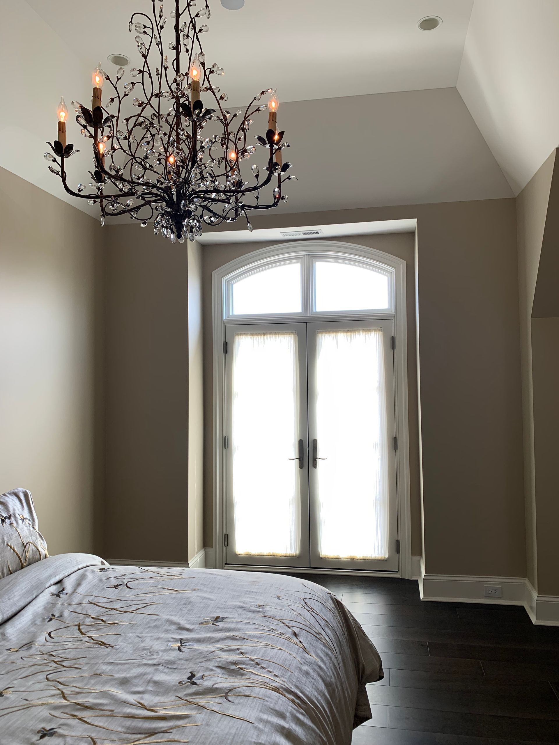 Bedroom with dark wood floor, tan walls, French doors, chandelier, and bed.