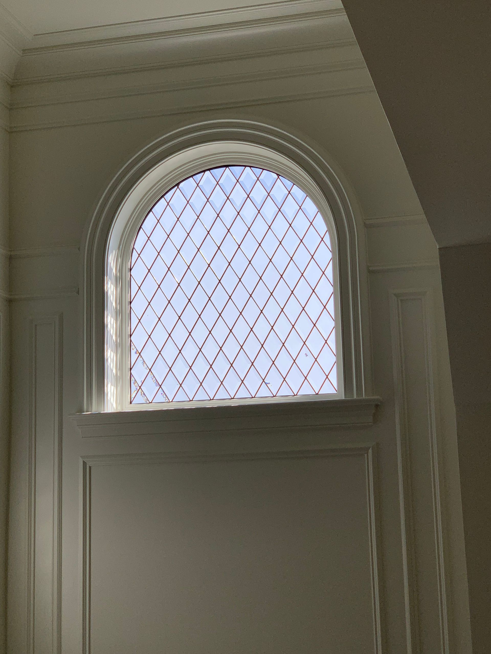 Arched window with diamond-patterned glass, set in white paneled wall. Blue sky visible.