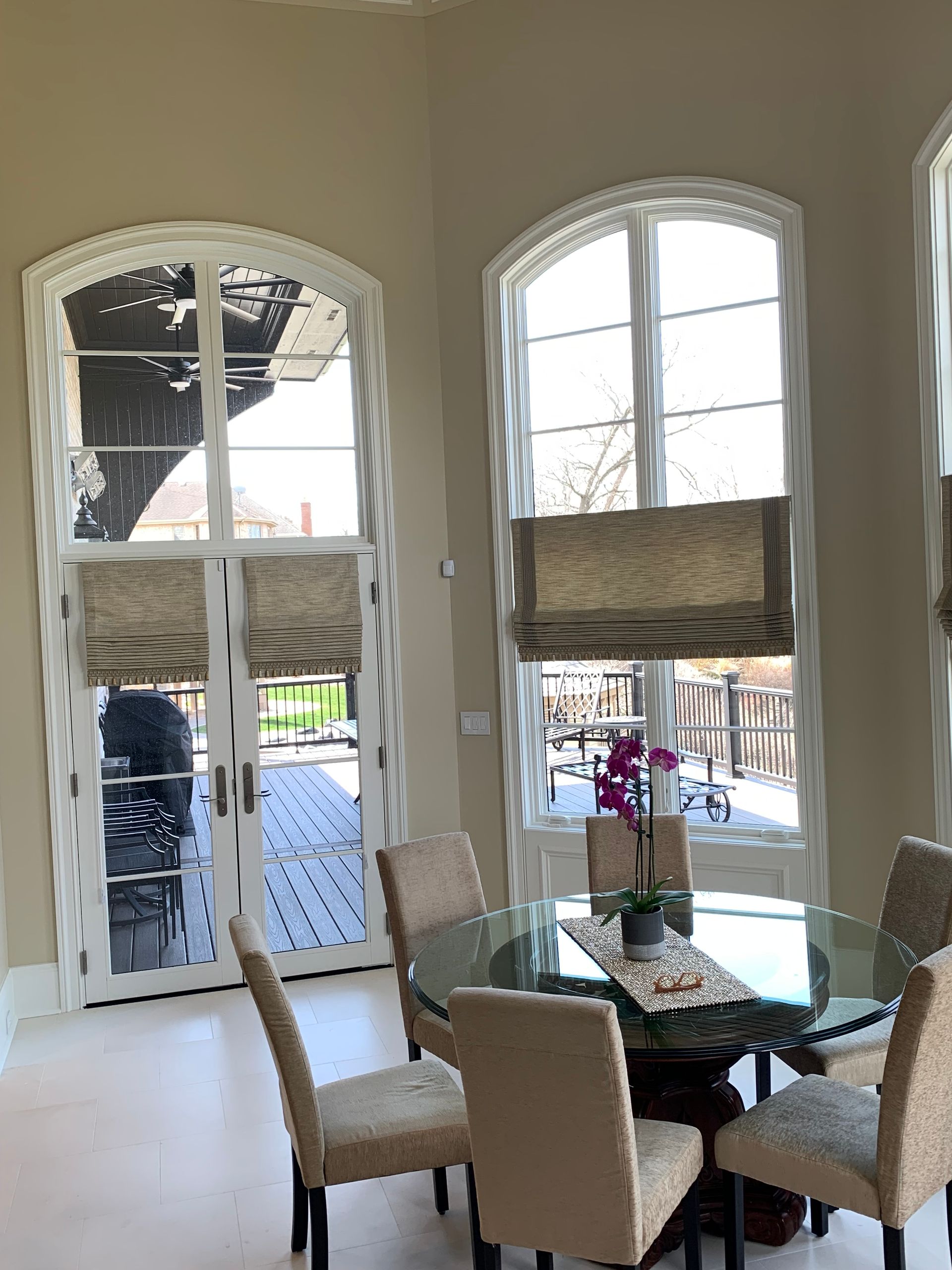 Dining room with a glass table and chairs, arched windows with shades, and a view of a deck.