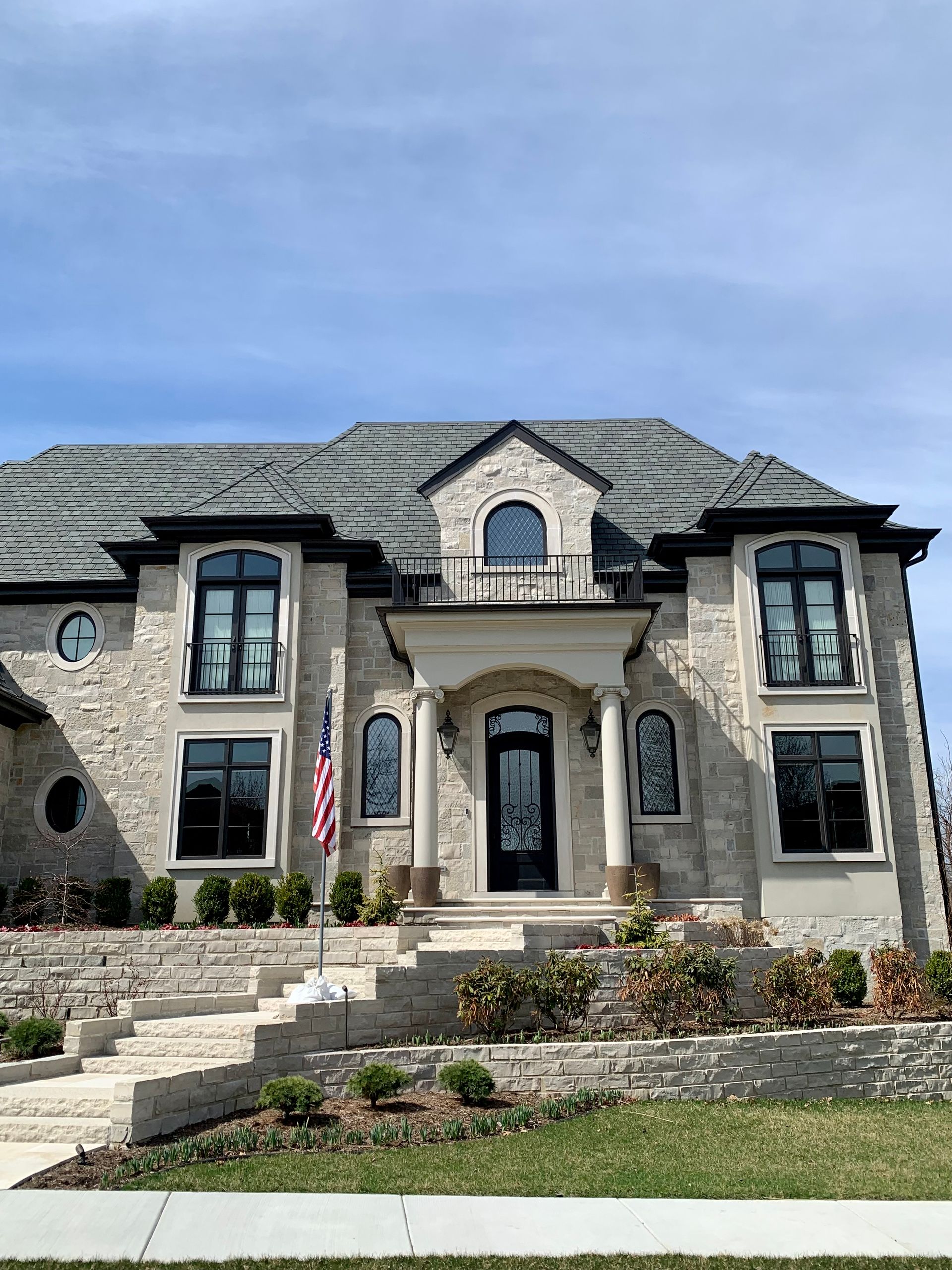 Stone mansion with black-framed windows and door, American flag, front steps, blue sky.