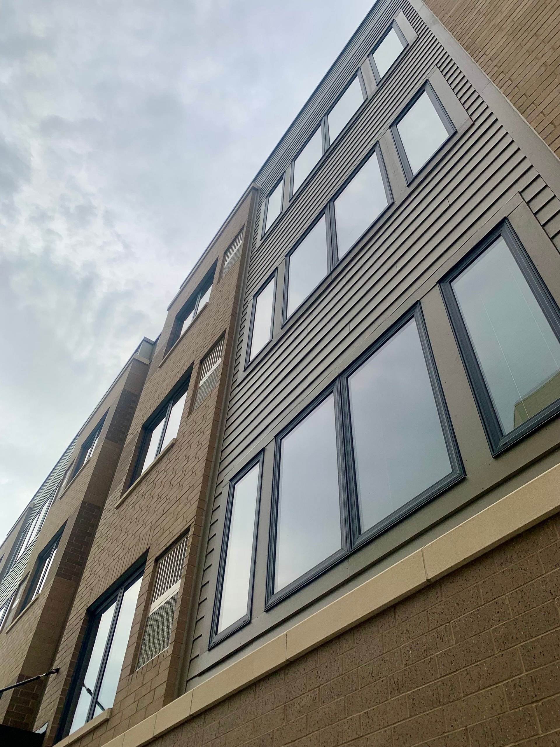 Low-angle view of a multi-story building with large windows and tan brick exterior under a cloudy sky.