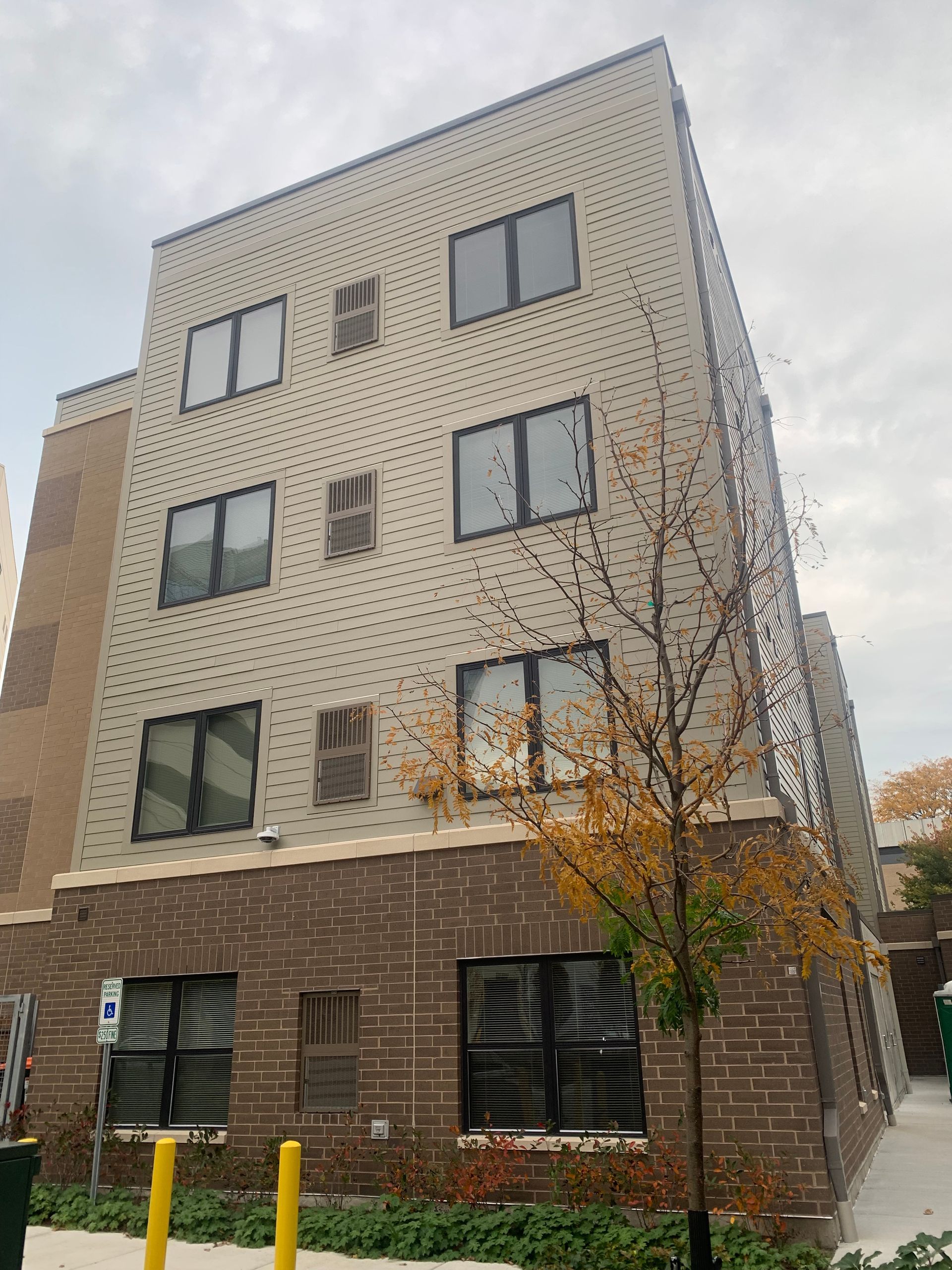 Multi-story brick building with windows, brown and tan facade, with small tree and overcast sky.