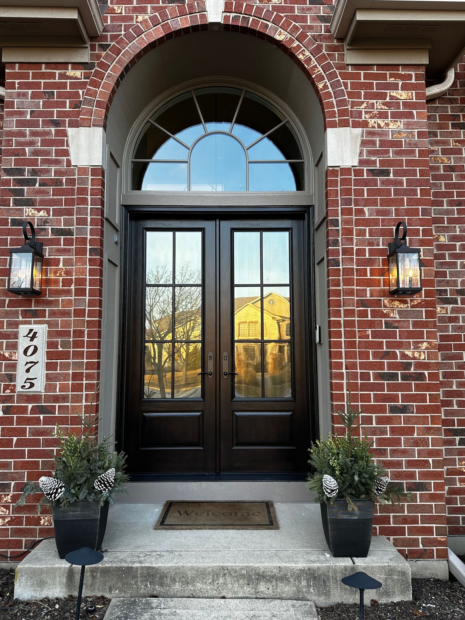 Brick home entrance with dark double doors, arched window, lanterns, planters, and stone steps.