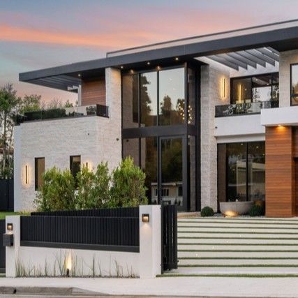 Modern two-story house with stone facade, wooden garage door, and black fence under a sunset sky.