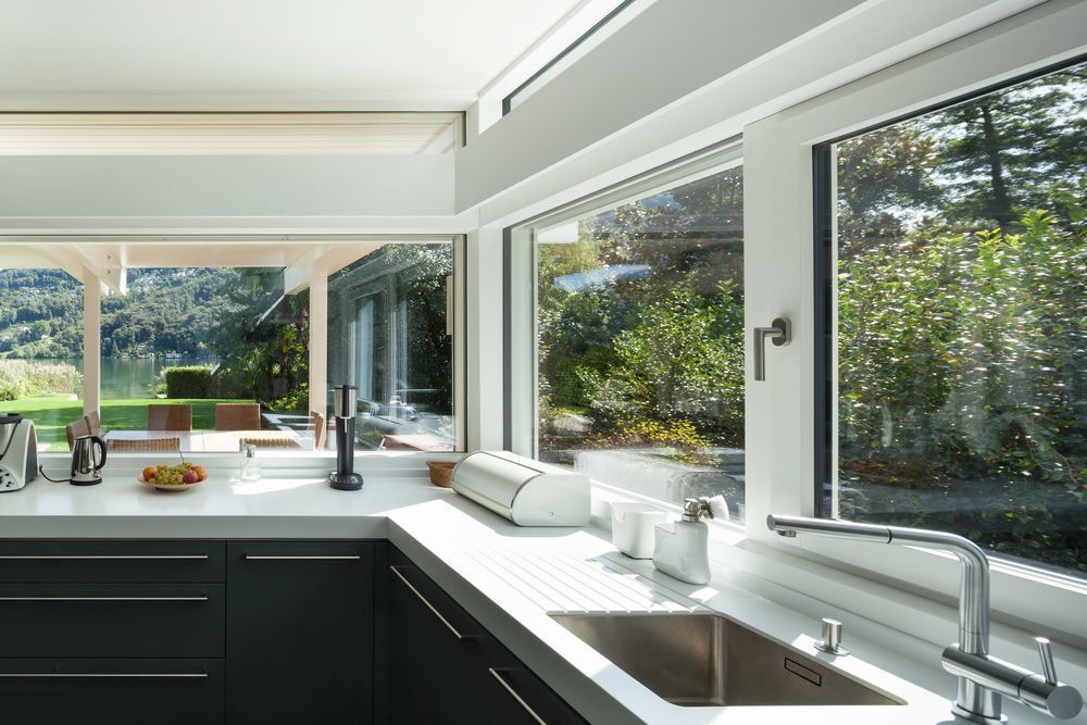 Modern kitchen with white countertops, dark cabinets, and large windows overlooking a backyard.