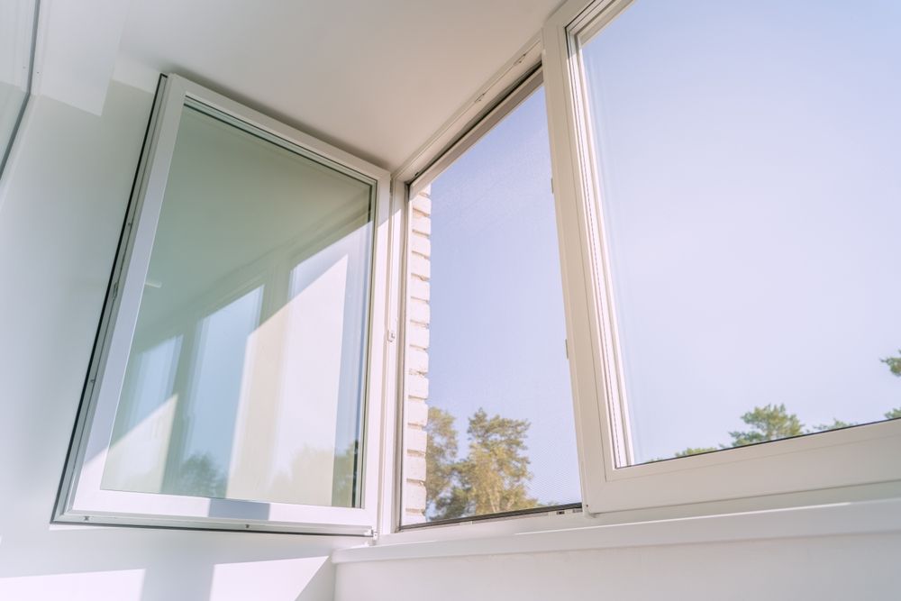 A view looking up at white window frames, with one open panel revealing a bright sky and trees.