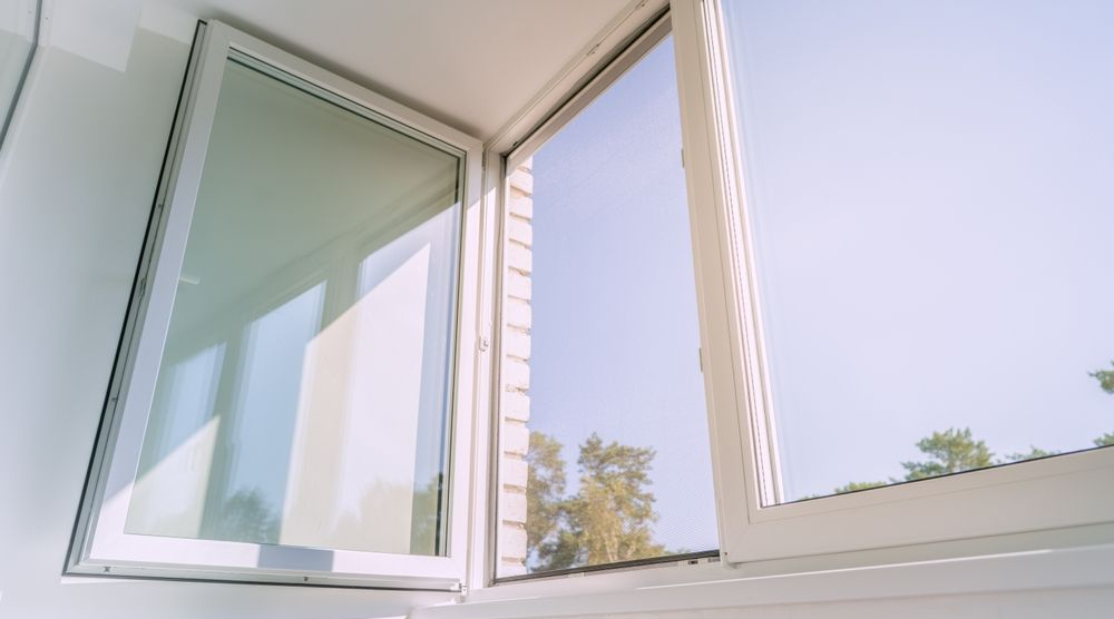 A partially open white-framed window looking out at trees against a bright, clear blue sky.