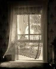 A black and white photo of a window with a lace curtain blowing in the wind.