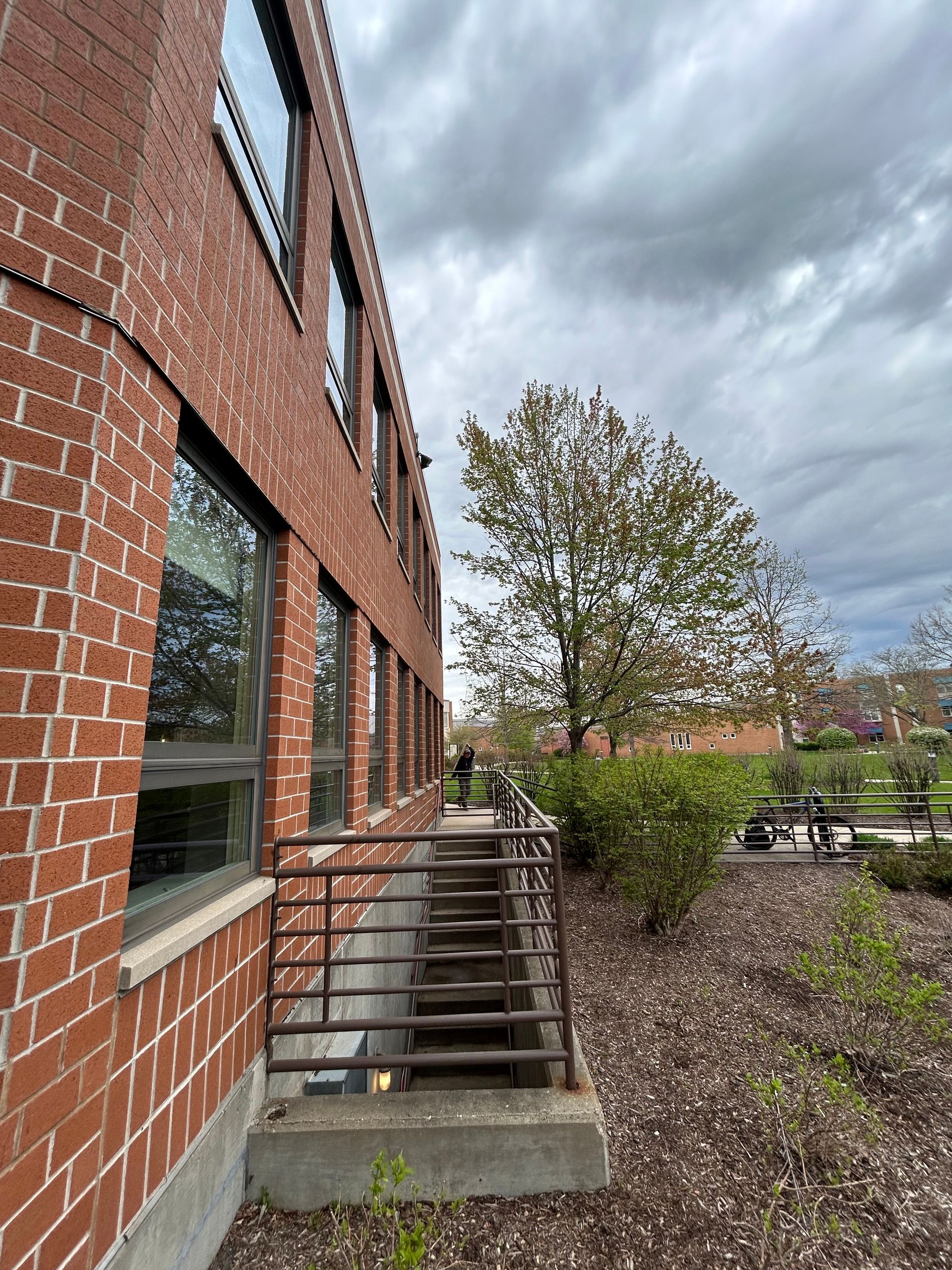Brick building with metal railing and stairs leading to an entrance, a tree, and cloudy sky.