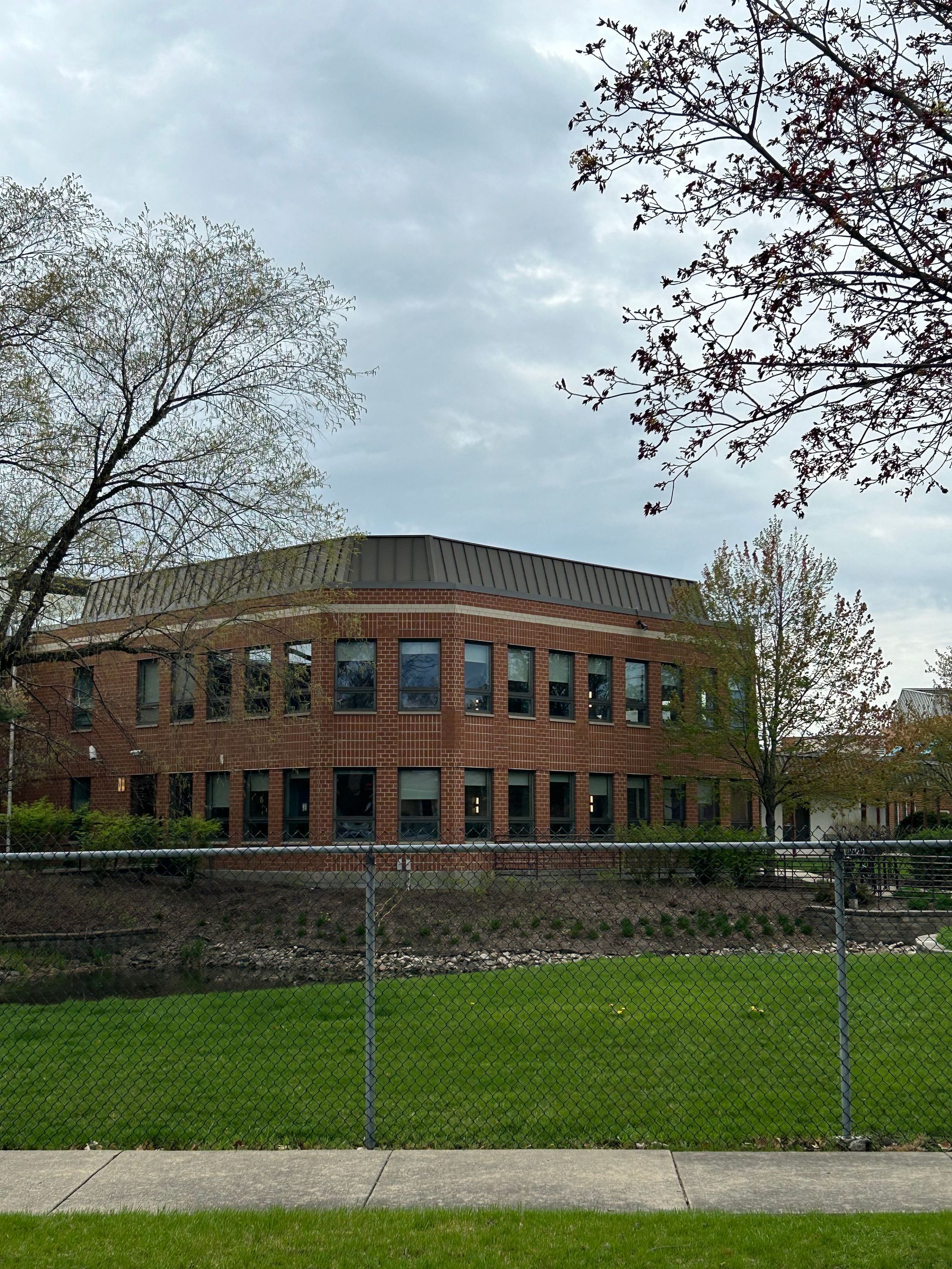 Brick building with many windows, behind a chain-link fence, under a cloudy sky. Trees in the foreground.