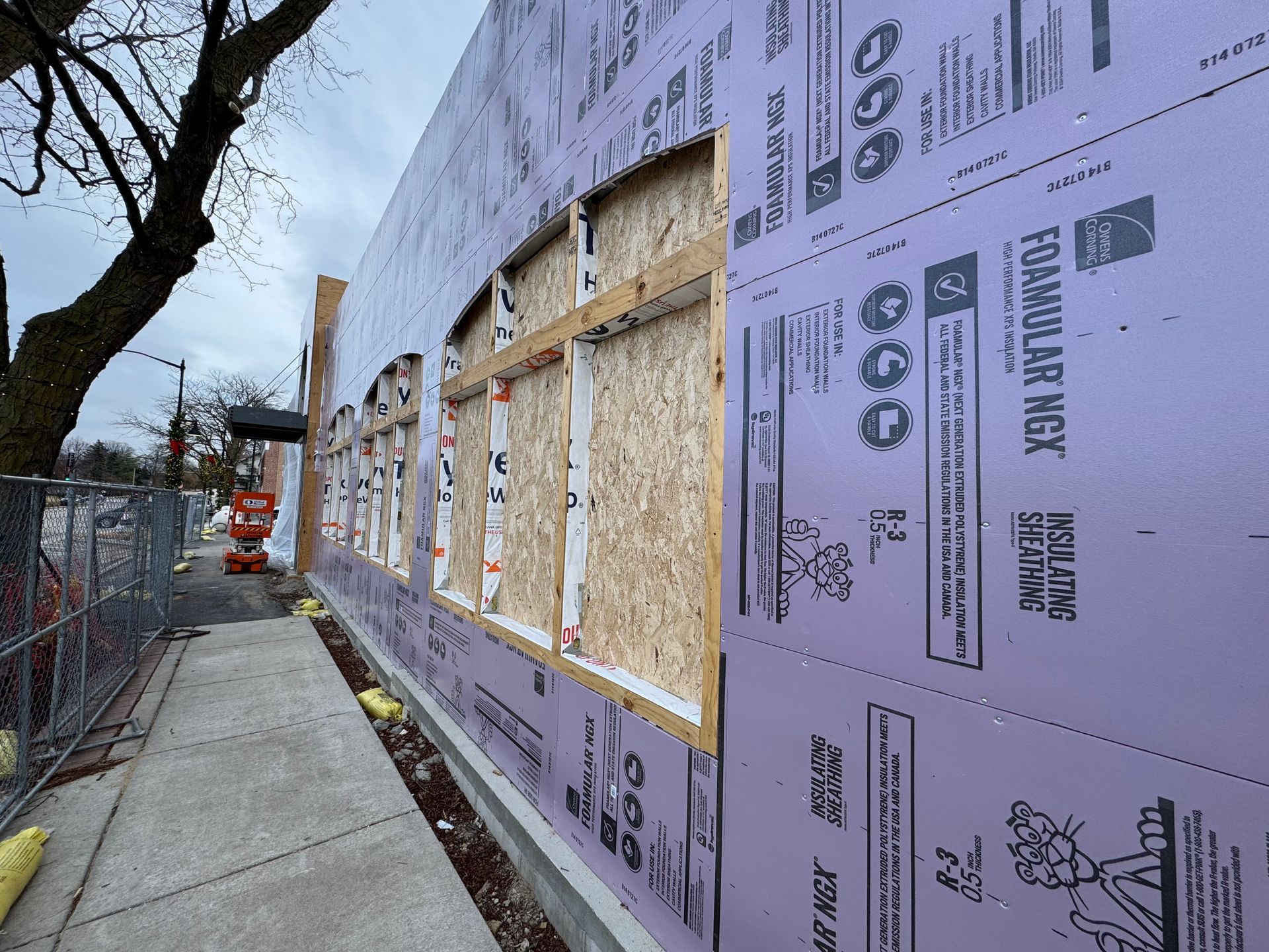 Exterior wall under construction with purple insulation, window frames, and a sidewalk.