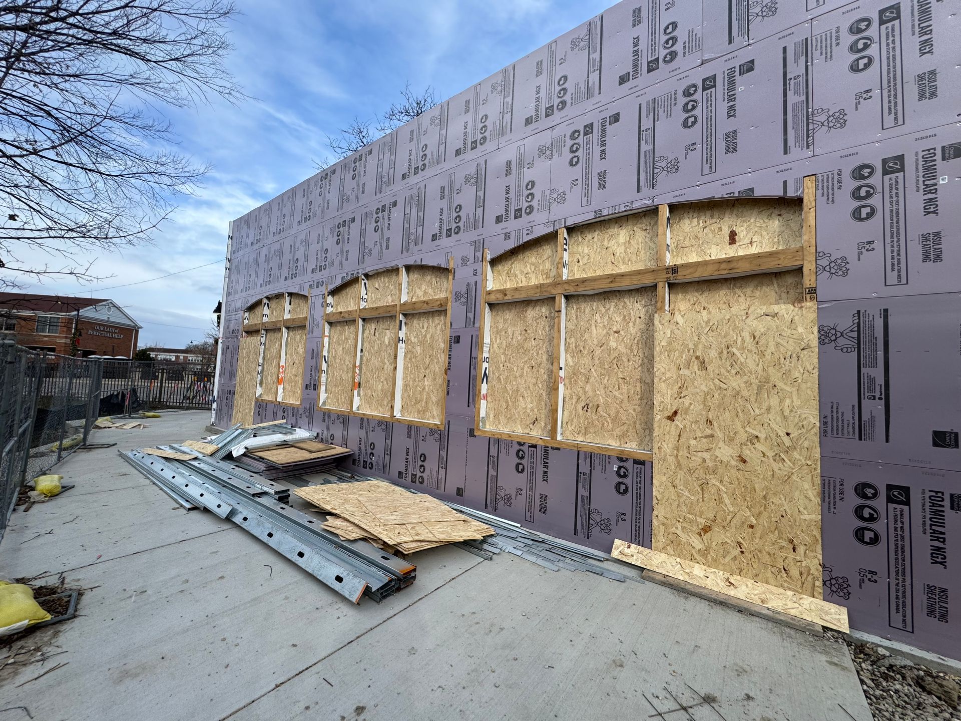Exterior of a building under construction, showing plywood-covered window openings, insulation, and a concrete foundation.