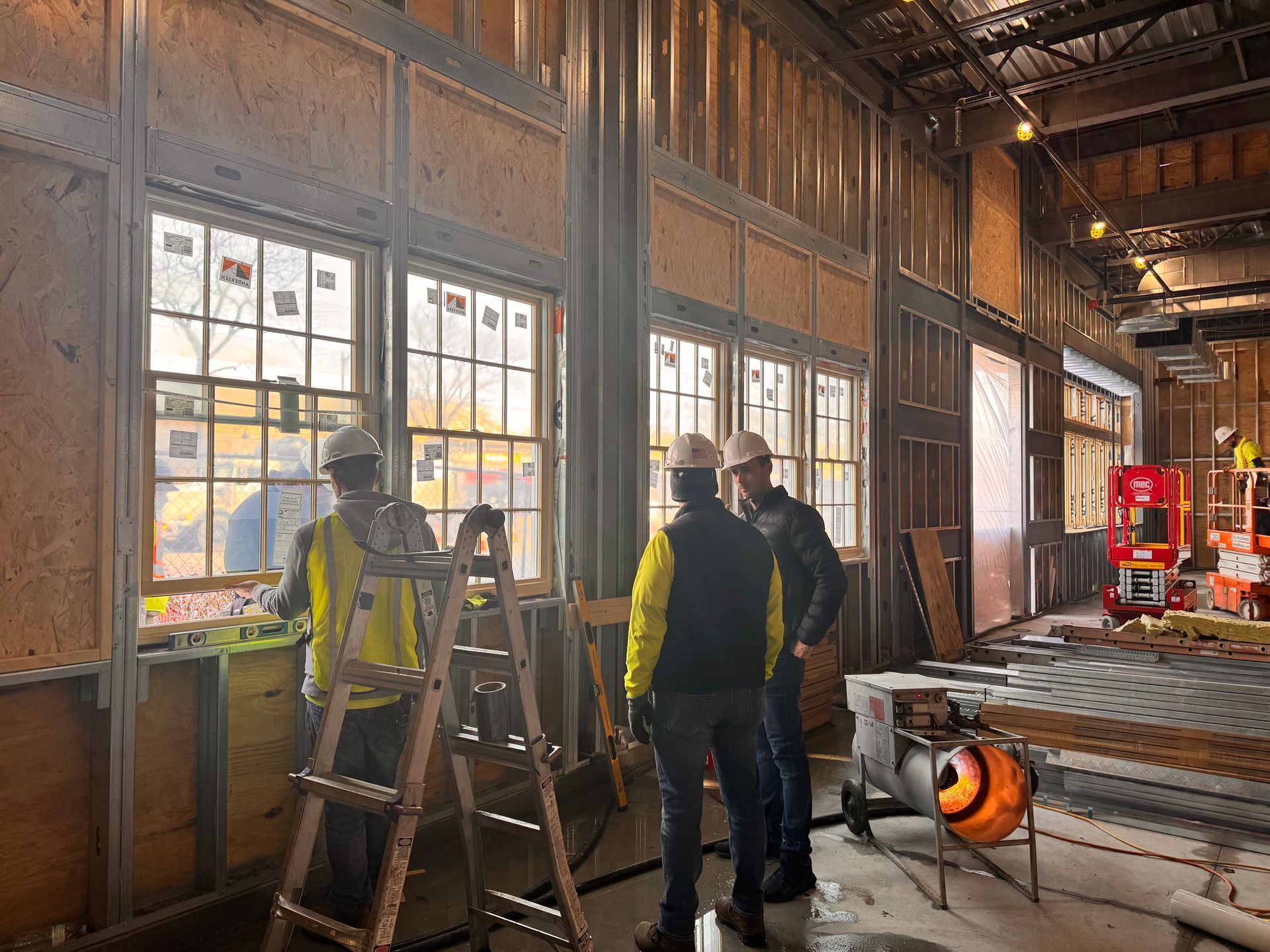 Construction workers inspect windows inside a building. One on ladder, wearing yellow vests, hard hats.