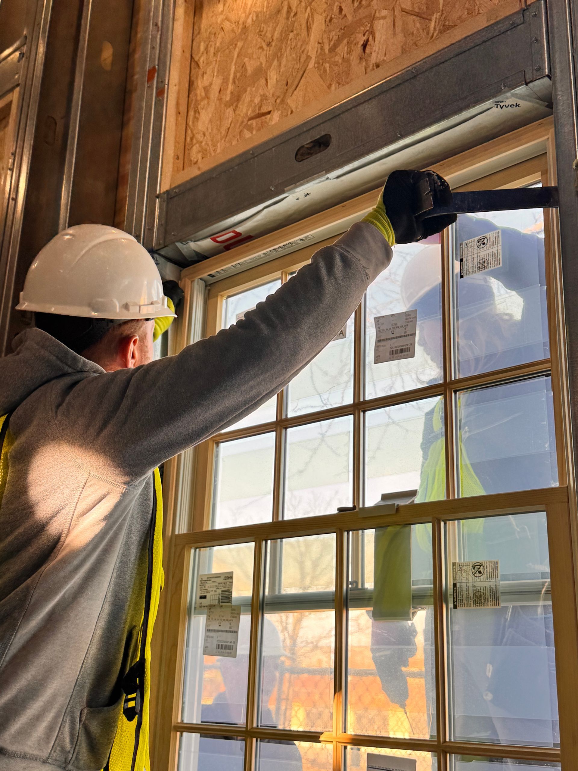 Construction worker installing a window, wearing a hard hat, safety vest, and gloves. The sun shines through.