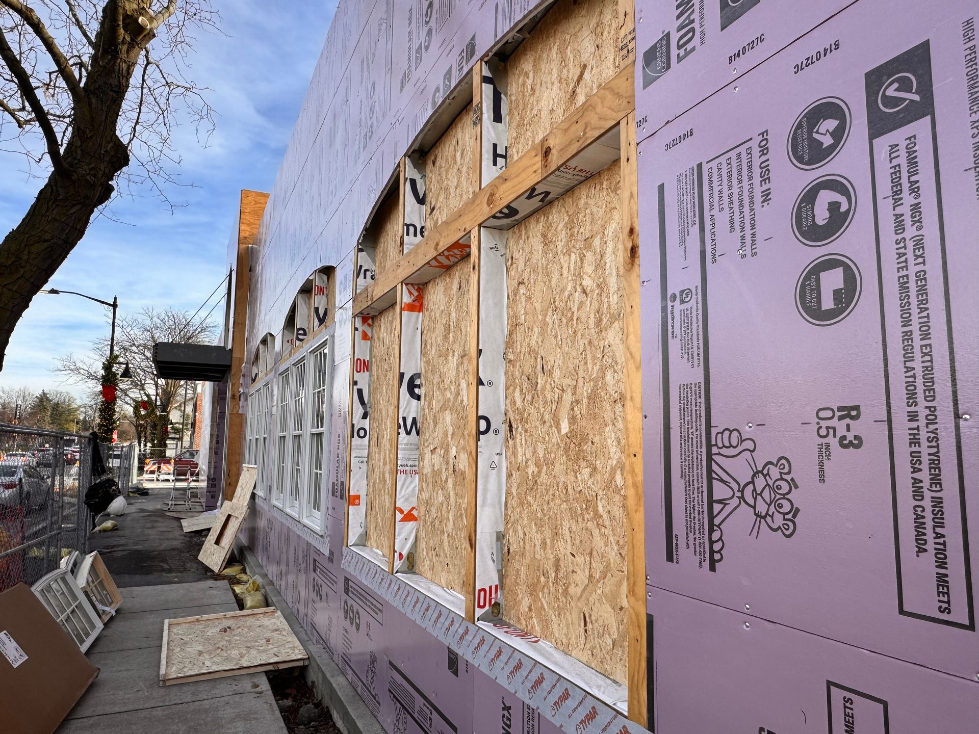 Exterior of building under construction. Windows are boarded up. Insulation is purple. Sidewalk visible.