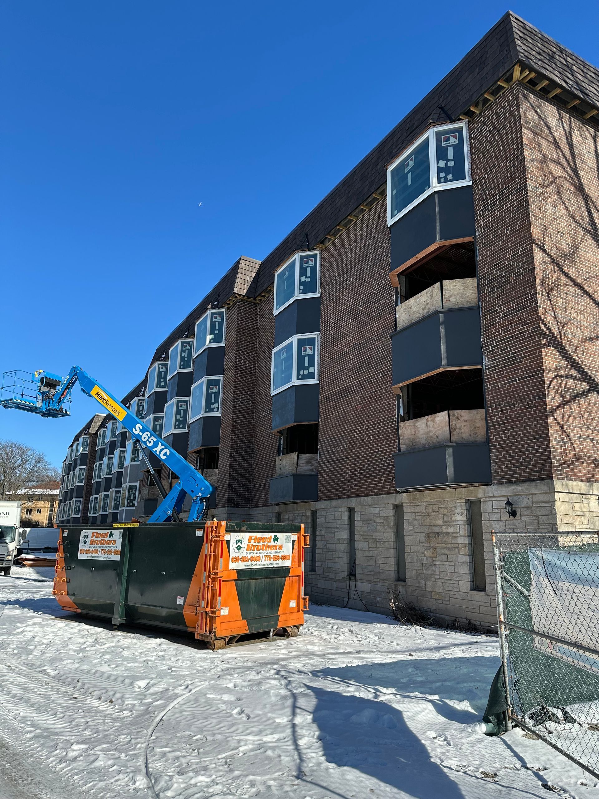 Building undergoing renovation; blue lift by dumpster; snow-covered ground.