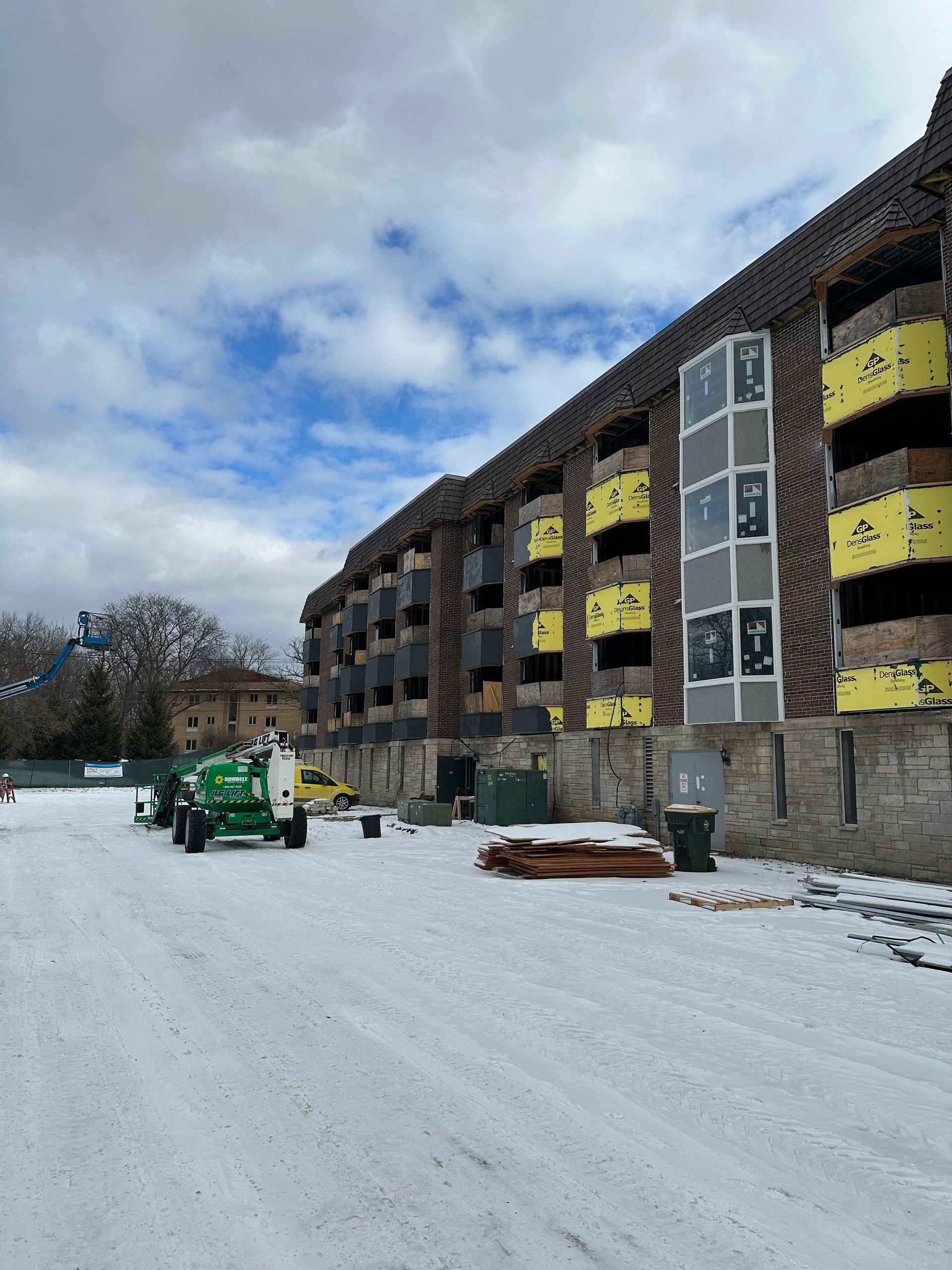 Building under construction on a snowy day. Brick facade, exposed insulation, construction equipment, and a cloudy sky.