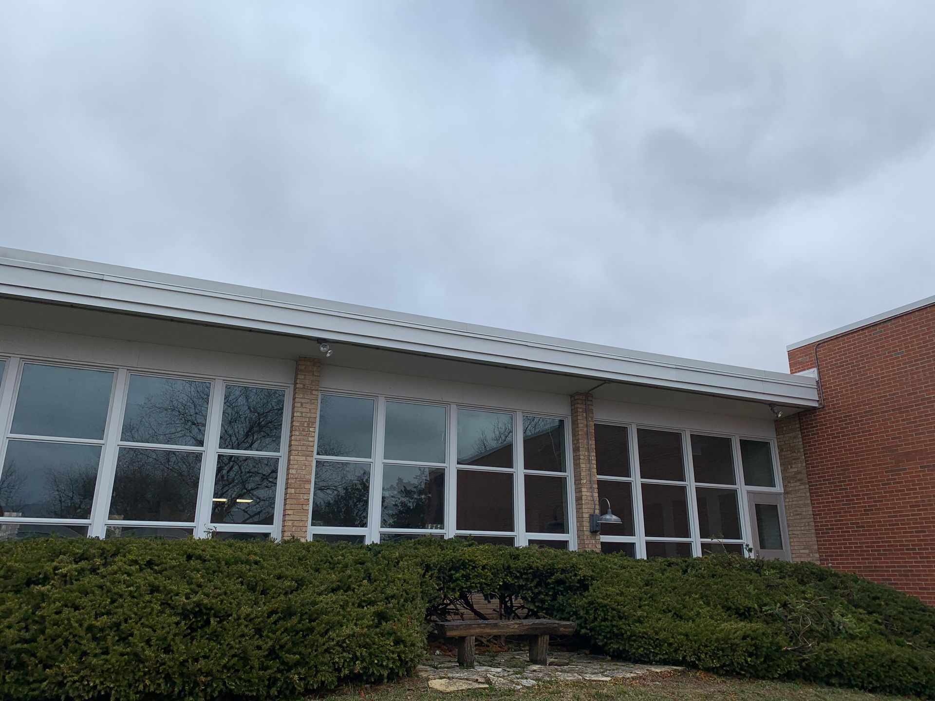 Building exterior with large windows, topped with a white roof. Brick wall to the right, shrubs below windows. Overcast sky.