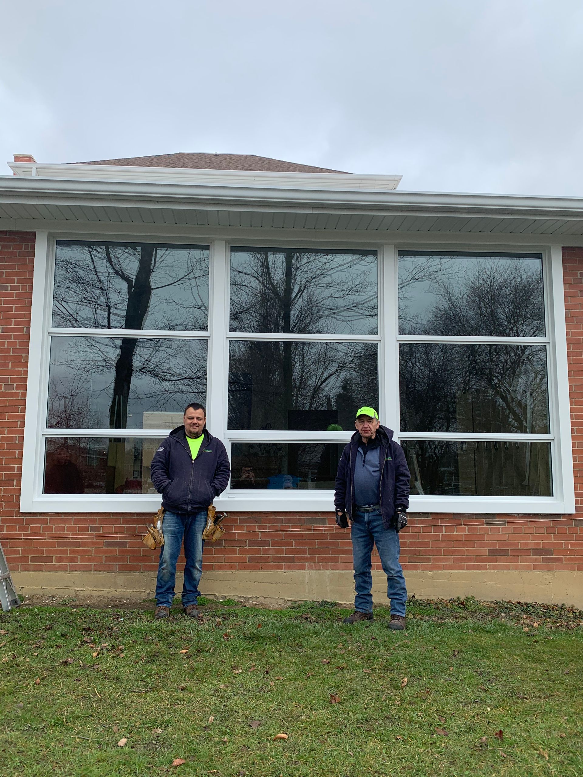 Two men stand in front of a large window on a brick building. Cloudy day, grass and trees.