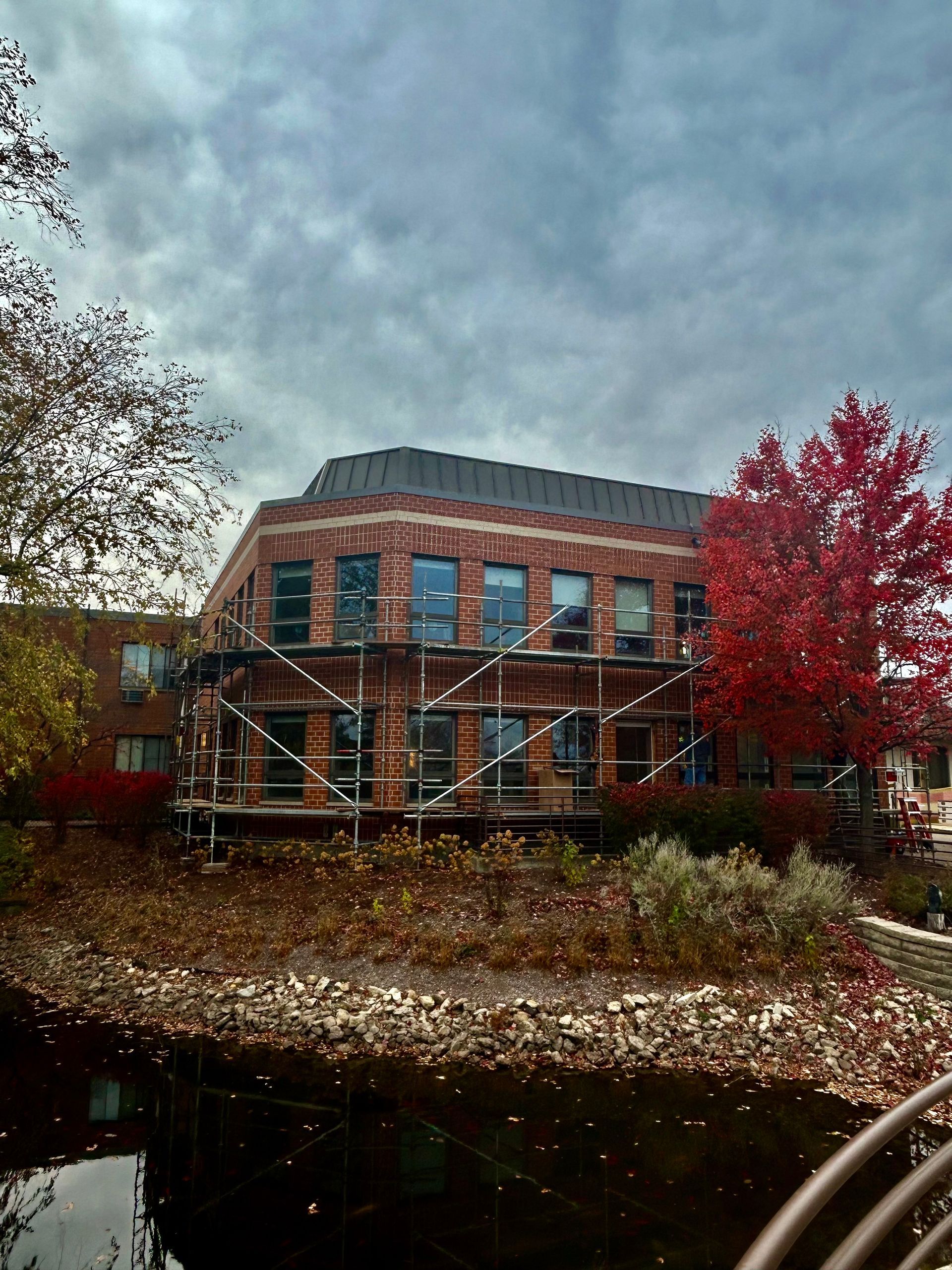 Brick building with scaffolding, near a river, autumn foliage and cloudy sky.