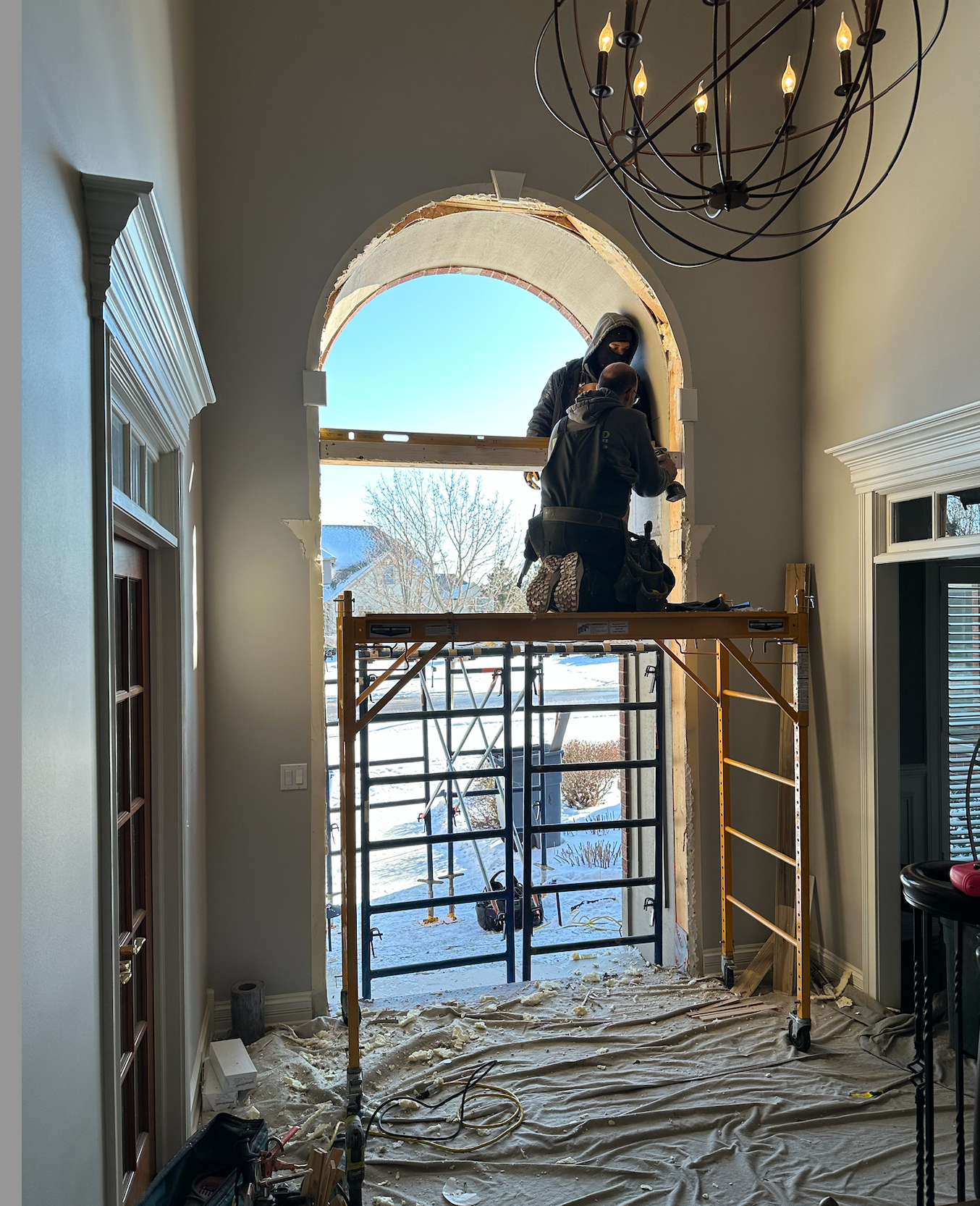 Two workers on scaffolding replacing a large arched window. Snow outside, chandelier above.