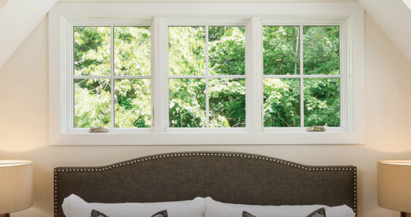A white-framed window with three panes looks out on lush green trees. A bed with a tufted headboard is below.
