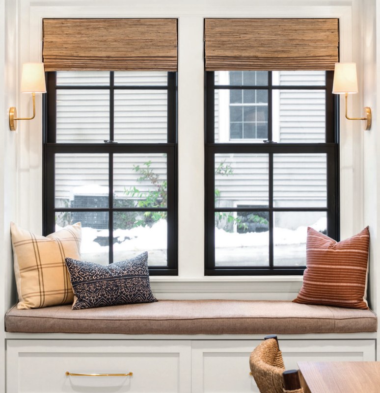 Two black-framed windows with woven shades, wall sconces, and a cushioned window seat with pillows.
