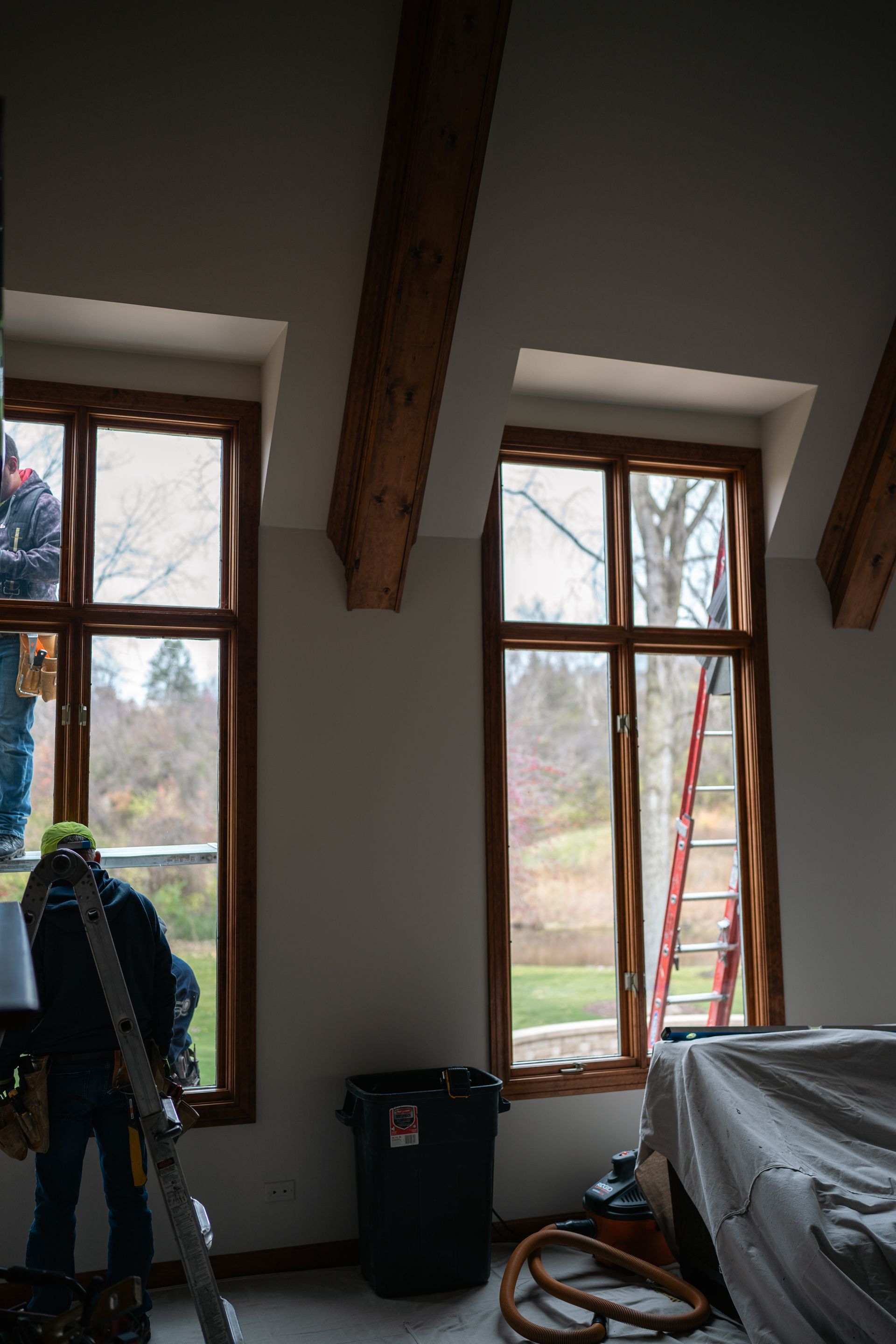 Workers near two brown-framed windows, looking out at greenery, with exposed wooden beams and a ladder.
