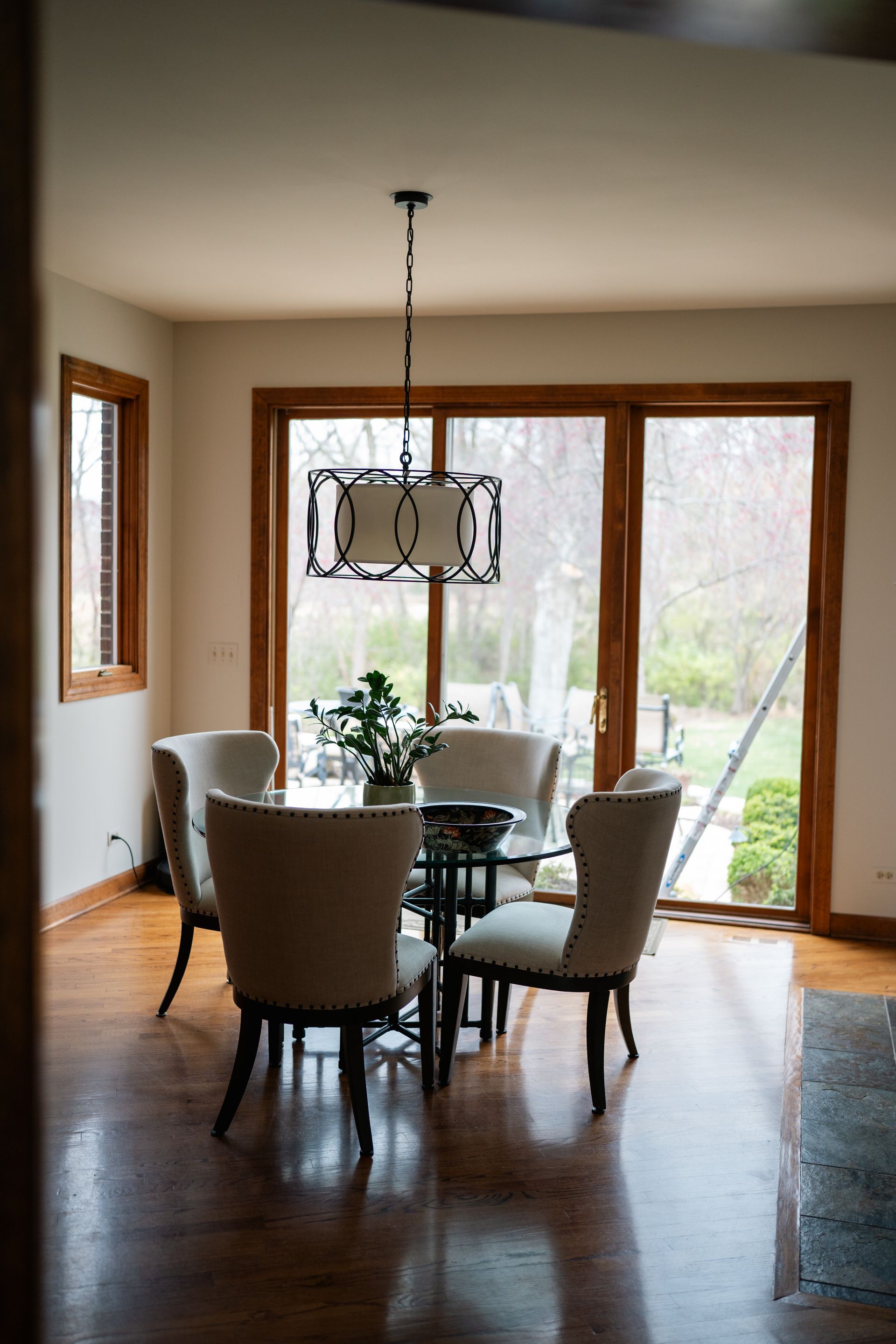 Dining room with round glass table, four upholstered chairs, pendant light, and large windows.