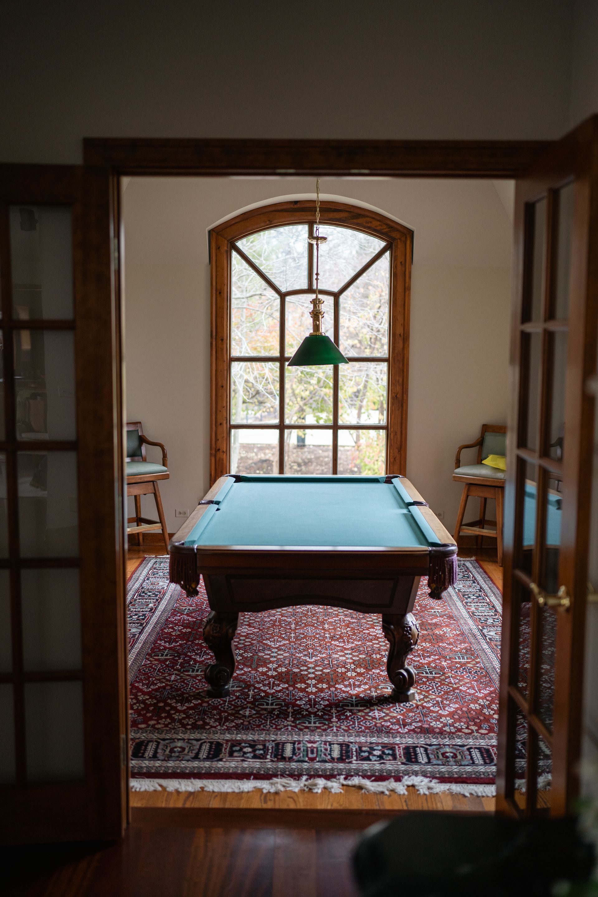 Pool table in a room framed by french doors; large arched window behind it, rug, chairs and green lamp.