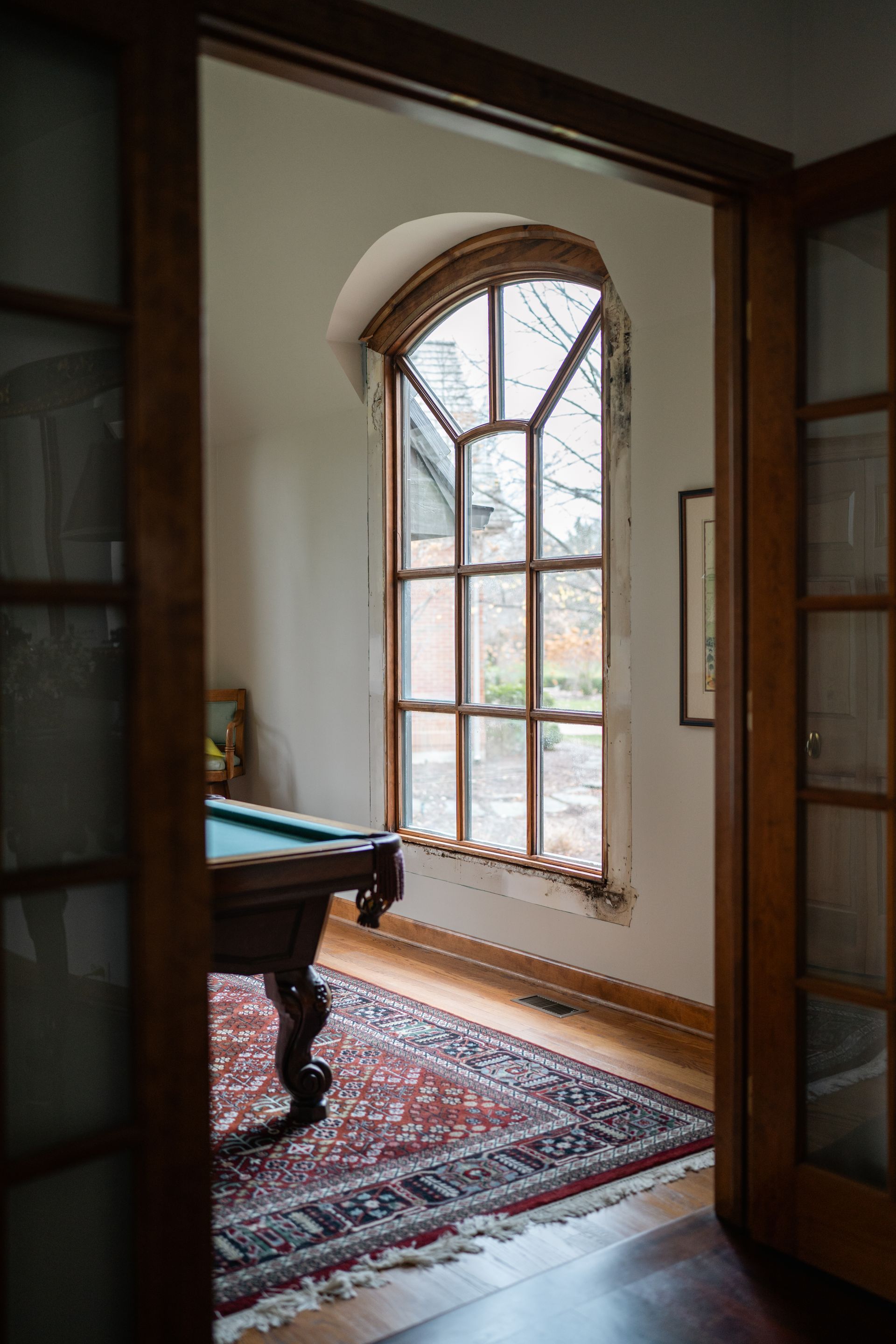 A pool table and rug in a room with a large arched window viewed through a doorway.