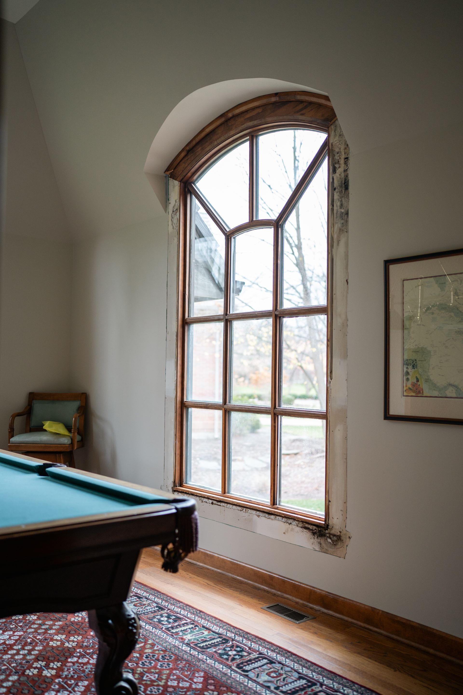 Pool table by large arched window; framed map on wall, chair in corner.