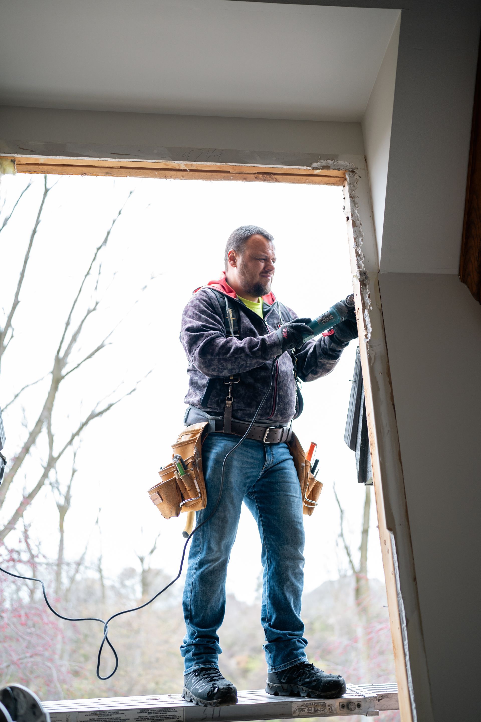 Man wearing tool belt, working on a window frame outdoors, holding a power tool.