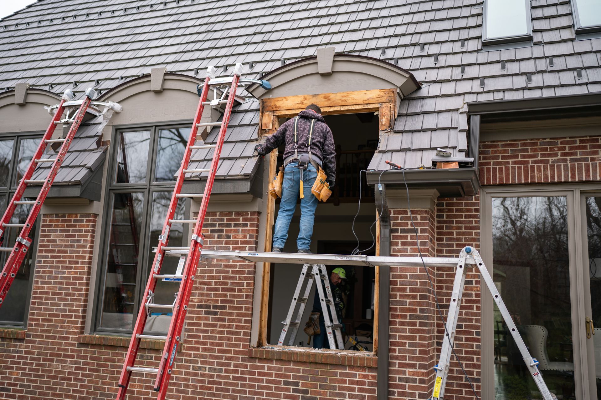 Construction worker on scaffold, replacing window in a brick building with shingle roof.