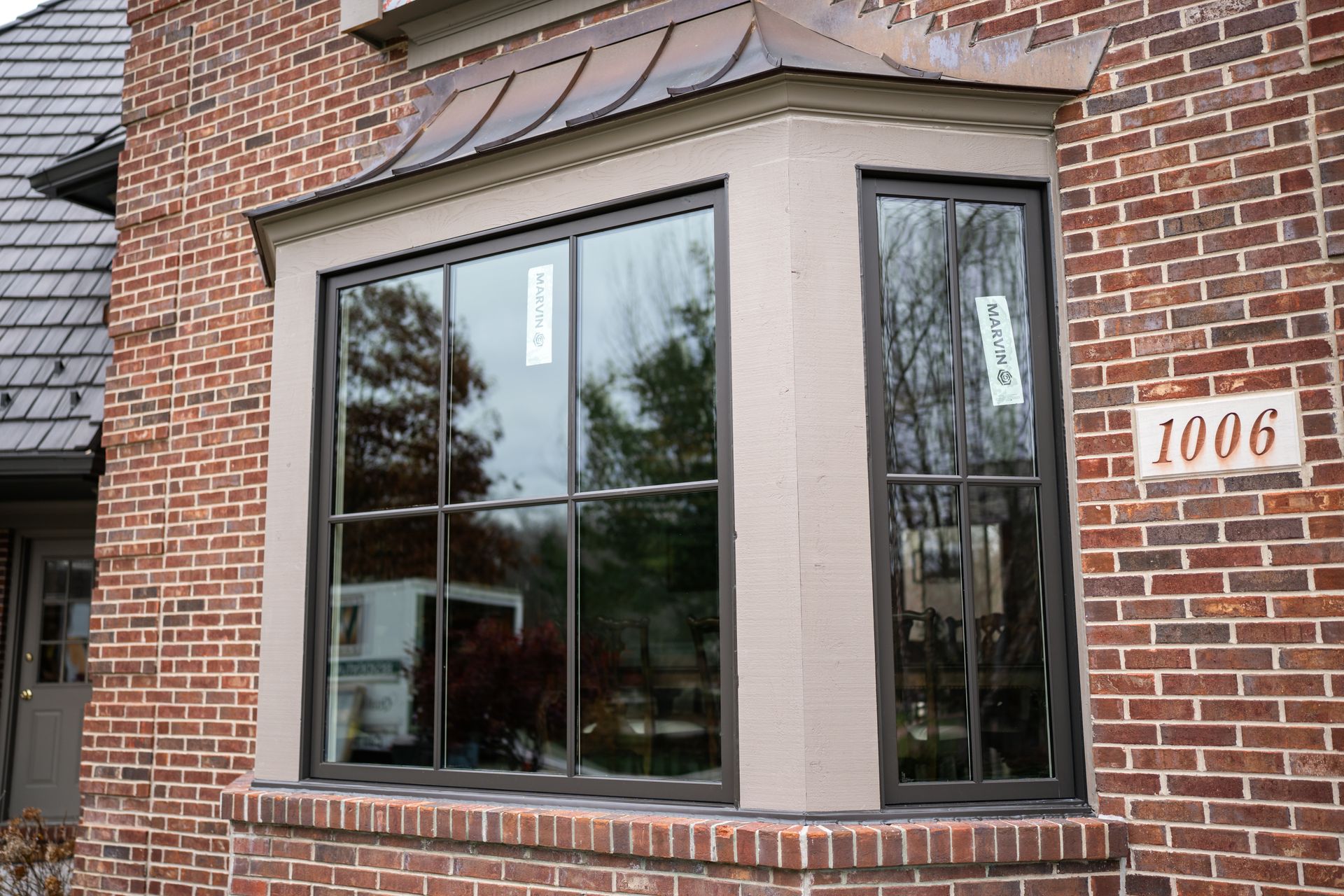 Bay window of a brick house with dark frames, featuring a copper-colored roof and the number 1006.