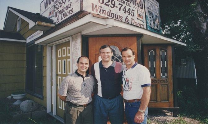 Three men are posing for a picture in front of a building that says windows and doors