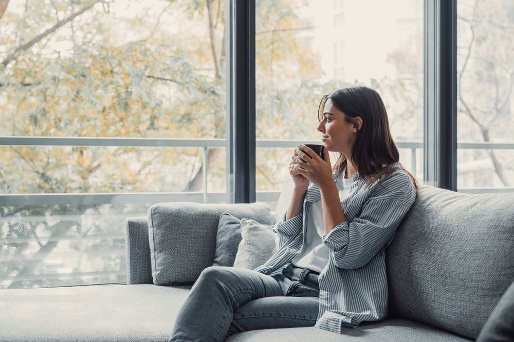 A woman is sitting on a couch drinking coffee and looking out the window.