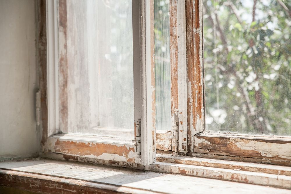 Close-up of an old, weathered window with peeling white paint and a blurry outdoor view.
