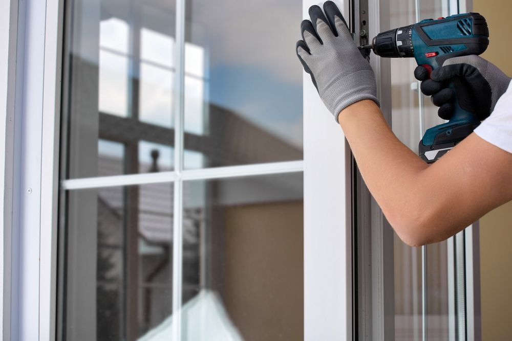 Person in gloves using a drill to install a white window frame.