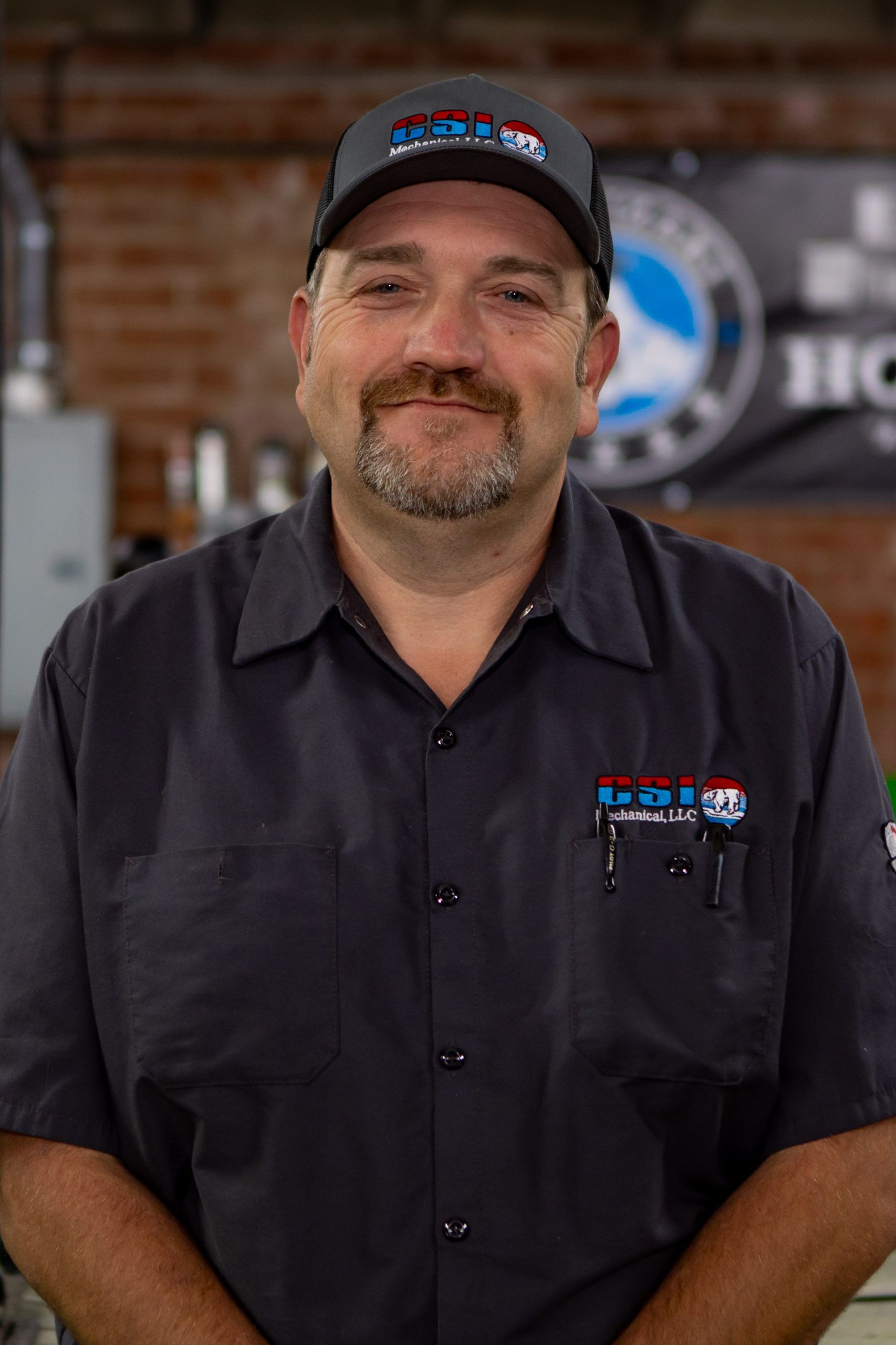A person wearing a gray work uniform and a branded baseball cap stands in front of a brick wall and logo signage.
