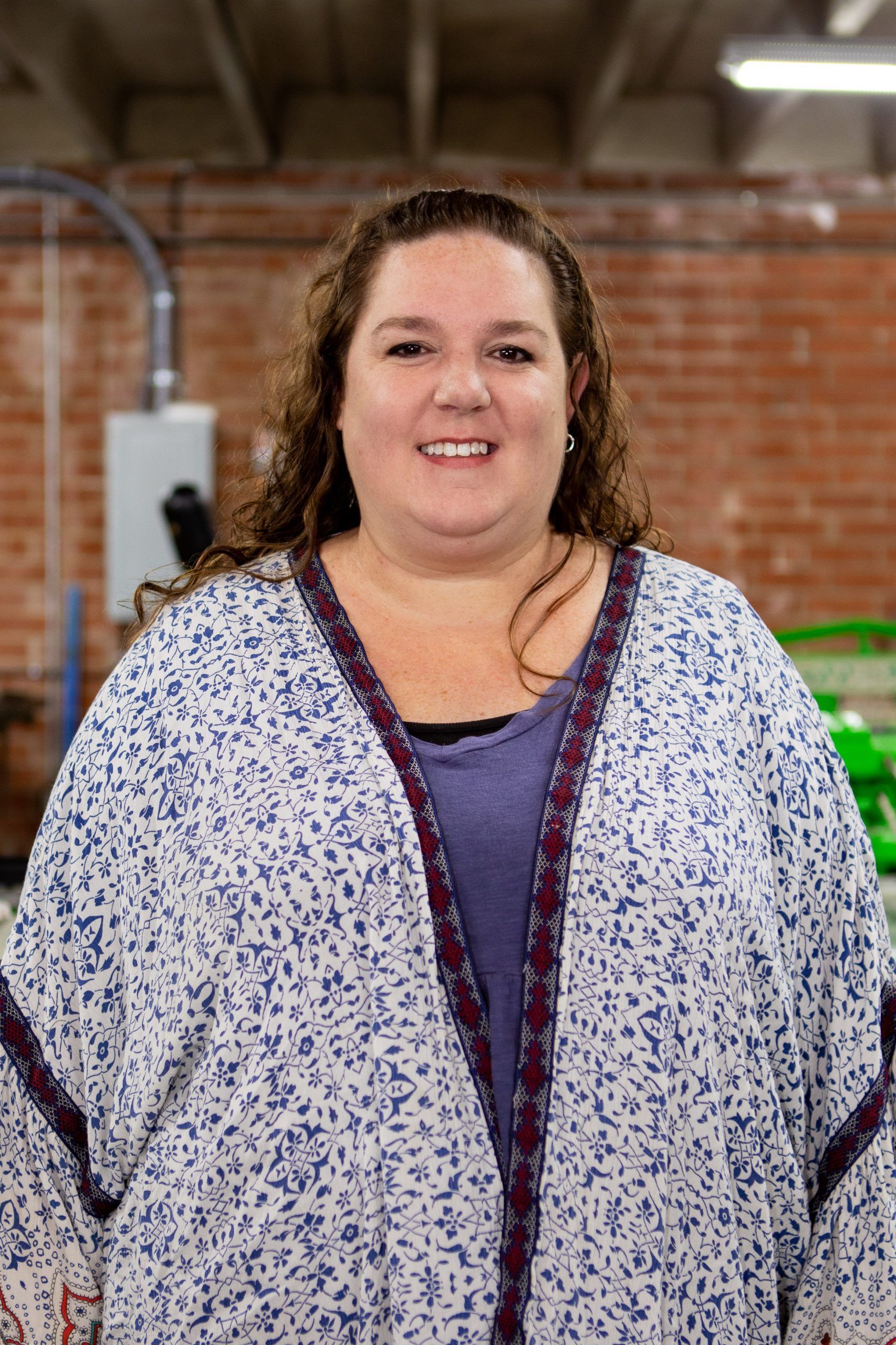 A smiling person with long curly brown hair wears a blue and white patterned kimono over a purple shirt.