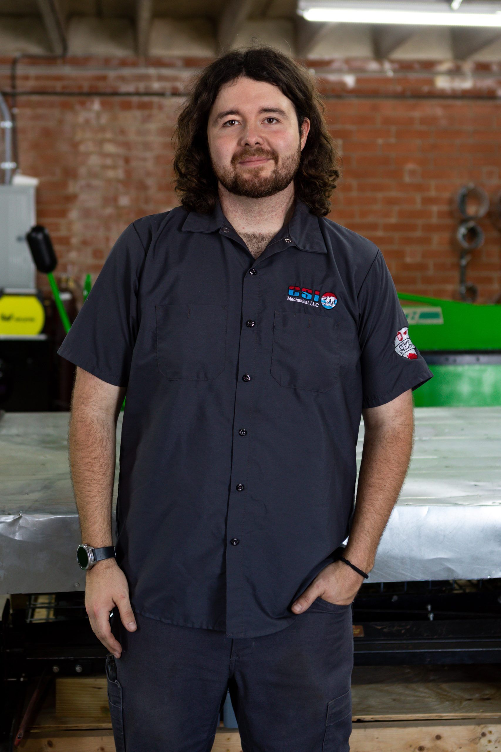 A person with dark, wavy hair wearing a grey work shirt and pants, standing in a workshop with a brick wall in the back.