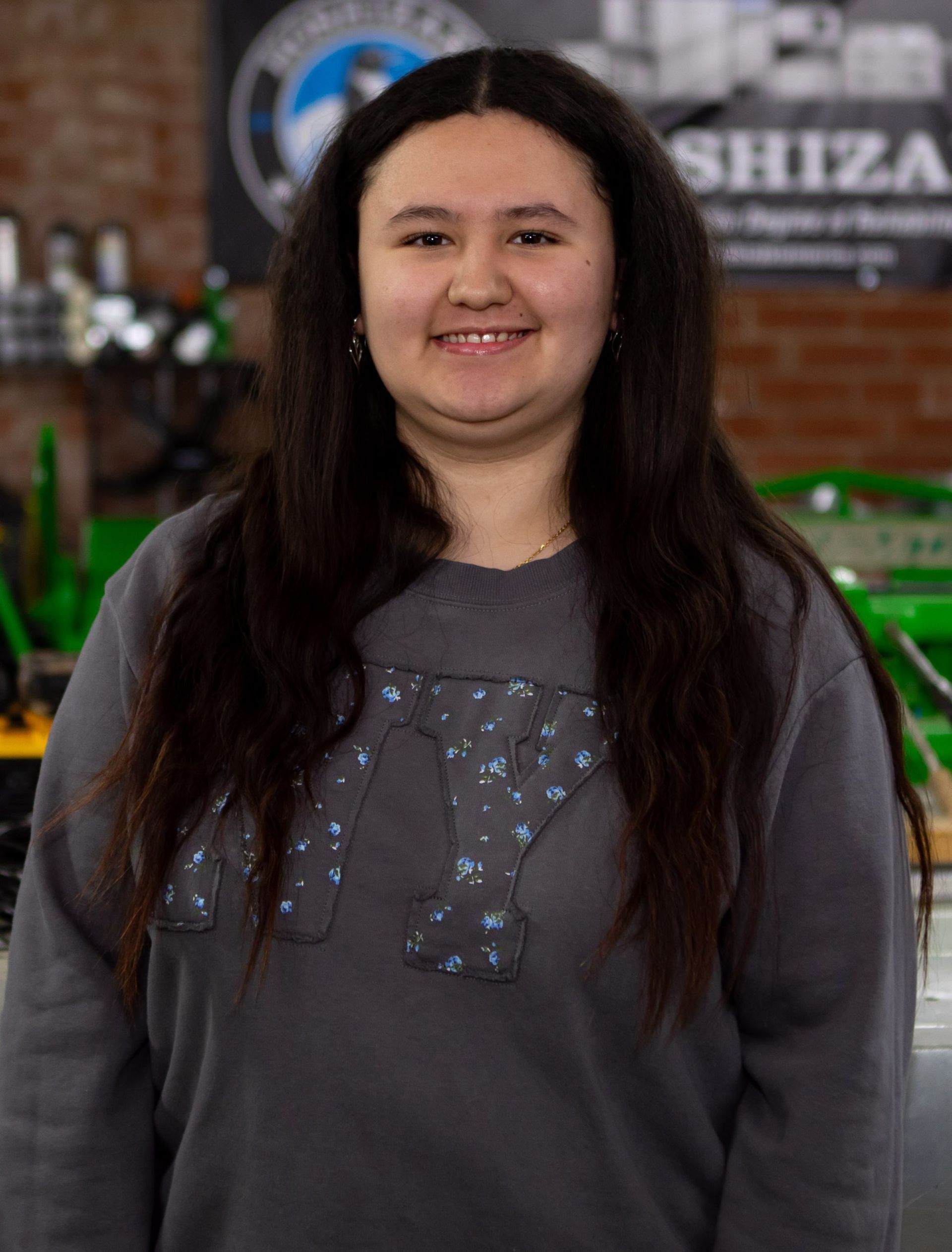 A person with long, dark hair wearing a gray sweatshirt with a patterned design, smiling in a workshop setting.