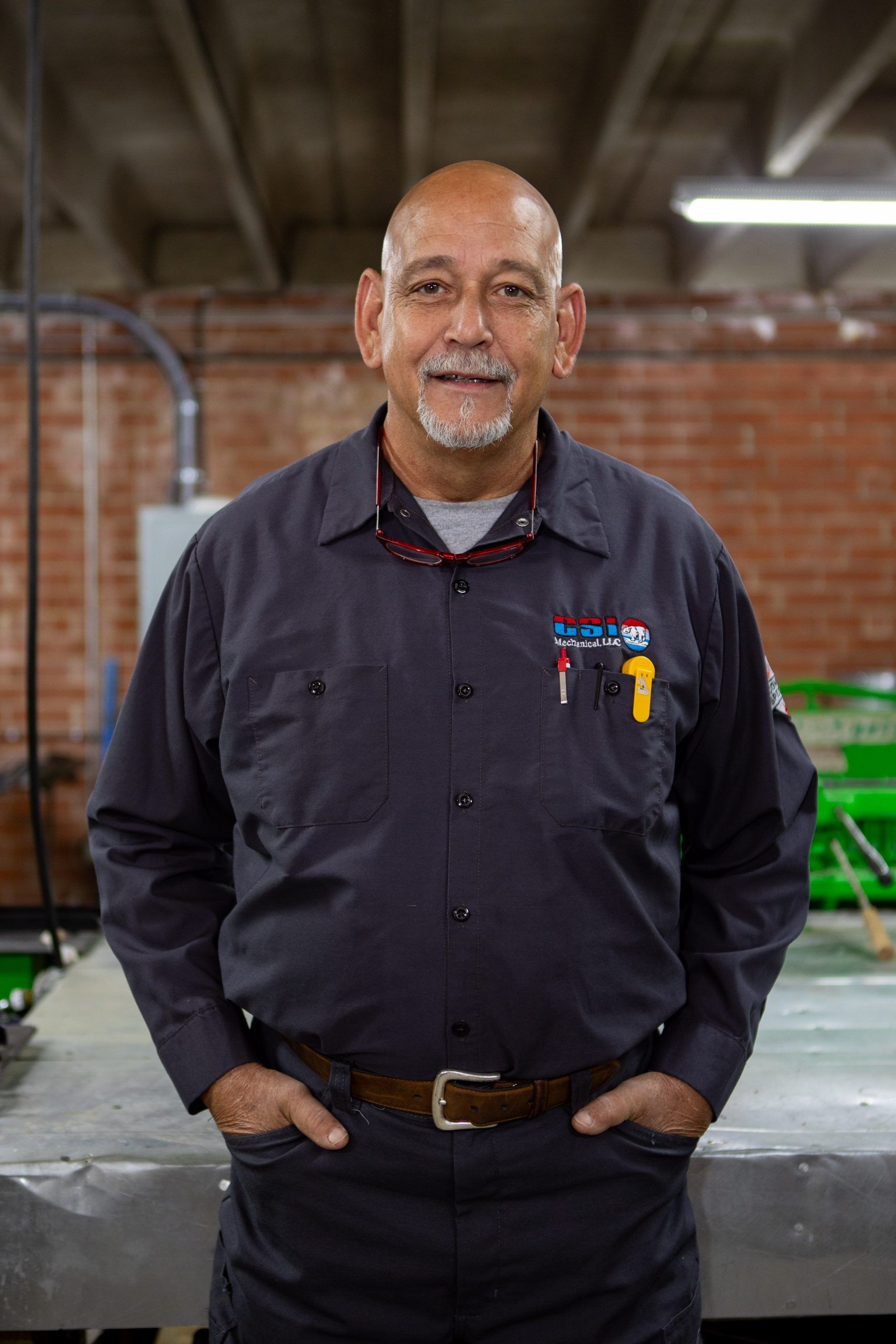 A person in a gray work shirt with pens in their pocket stands with hands in pockets against a brick wall background.