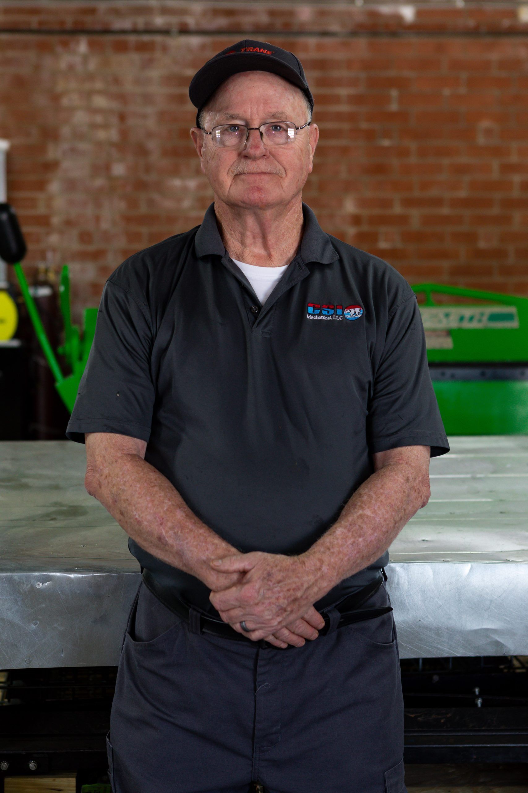 A person in a gray polo, dark cap, and glasses stands with hands clasped in front of them in an industrial workshop.