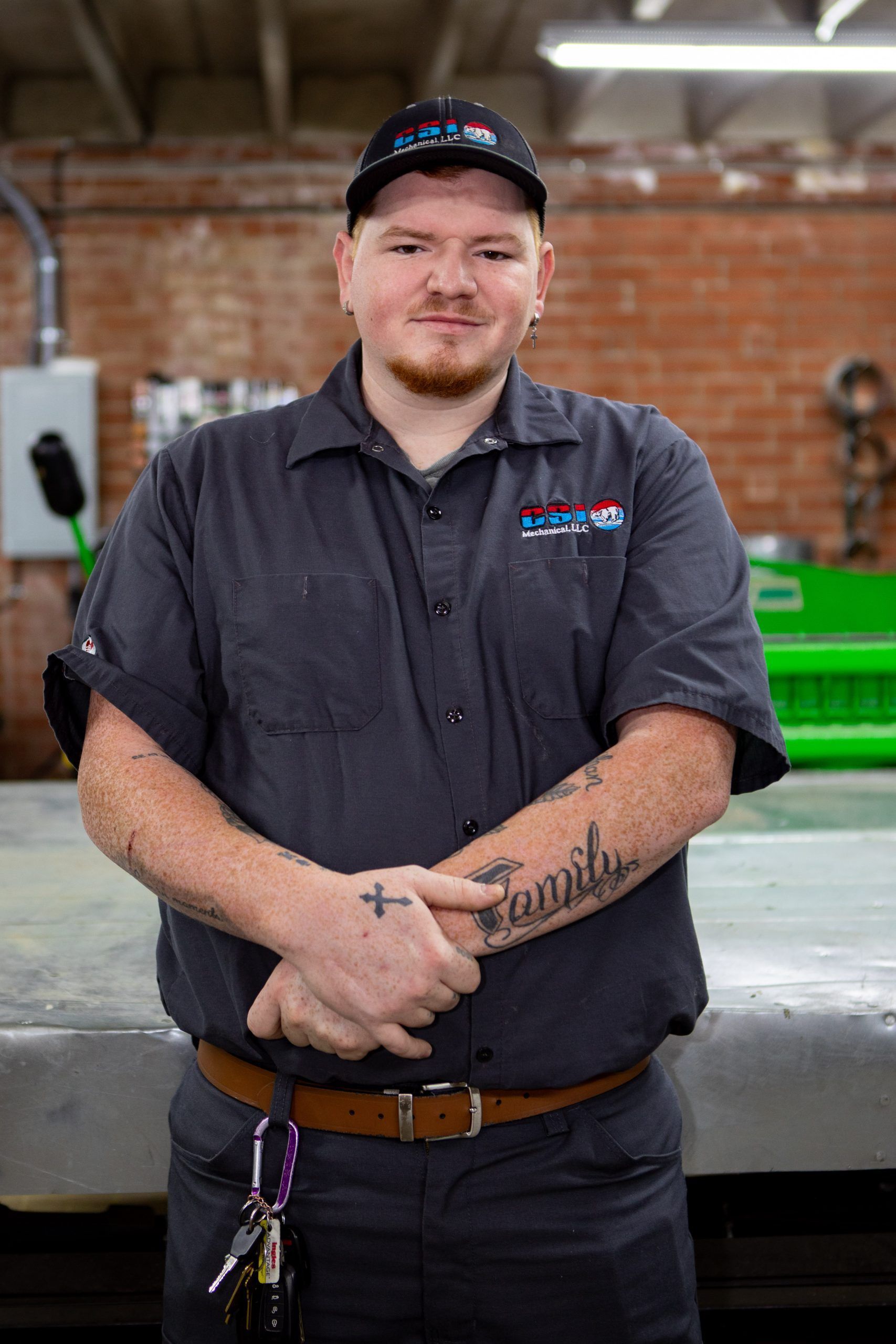 A worker in a gray uniform and cap stands with arms crossed in front of a brick wall in a workshop.