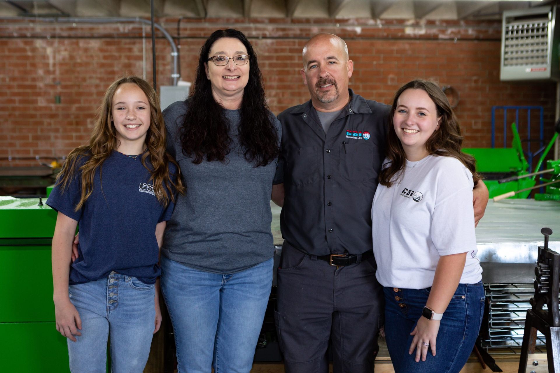 Four people stand side-by-side and smile in a brick-walled workspace with industrial green machinery in the background.