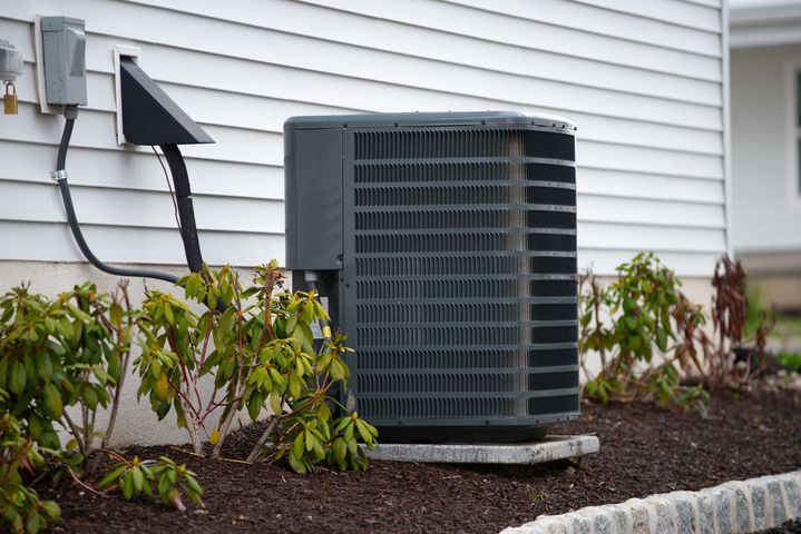 A gray outdoor air conditioning condenser unit sits on a concrete pad next to a white-sided house with landscaping.